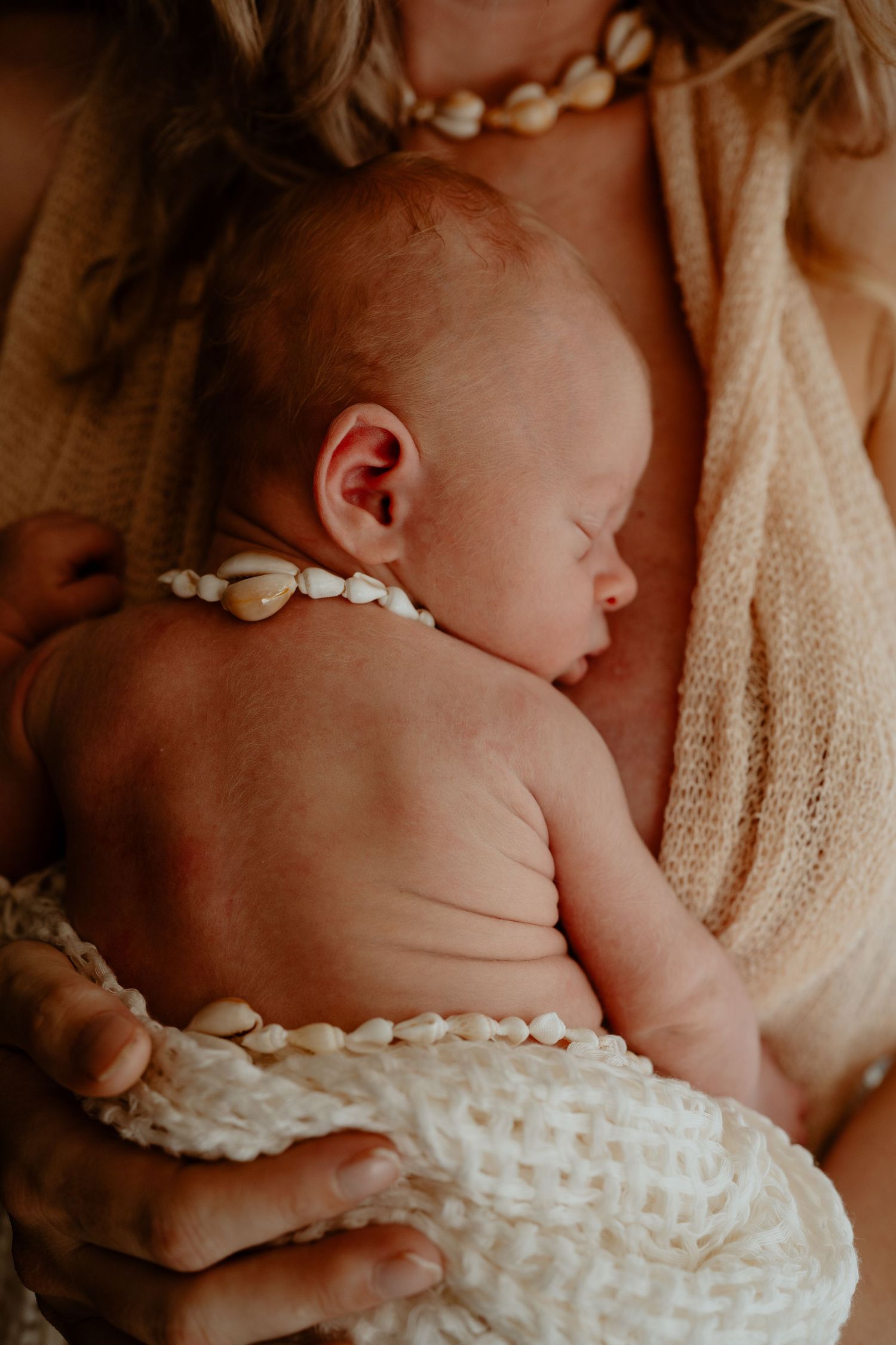 Close-up of sleeping newborn baby wearing cowrie shell necklace cradled against mothers chest.