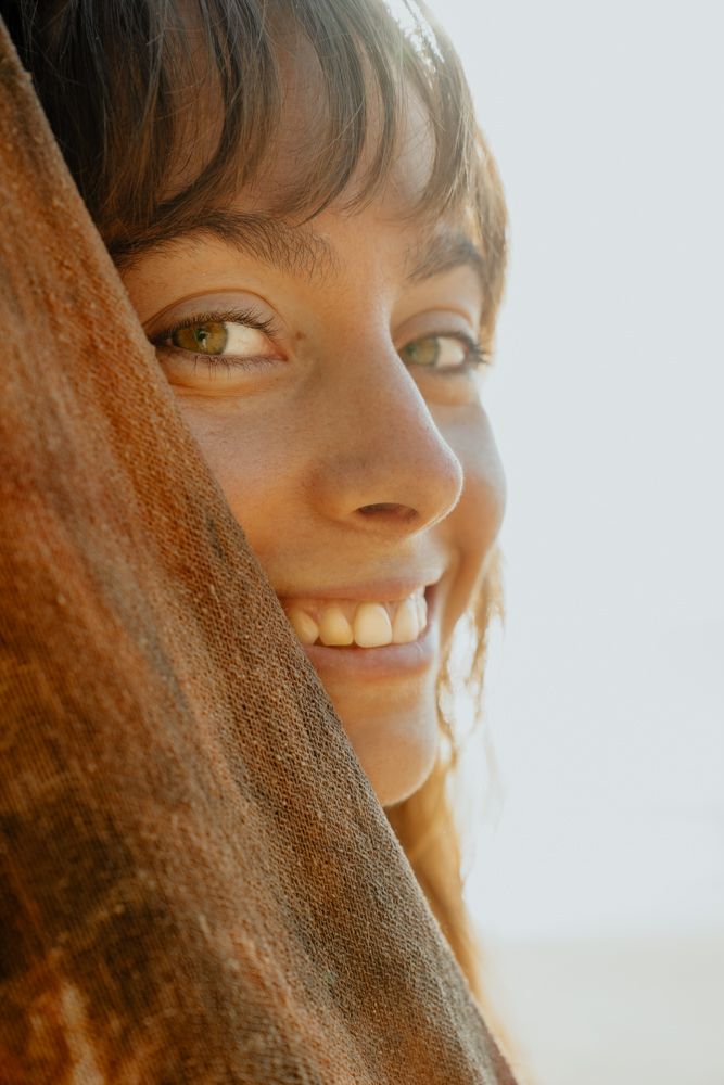 Close-up portrait of smiling woman with green eyes peeking behind brown fabric.