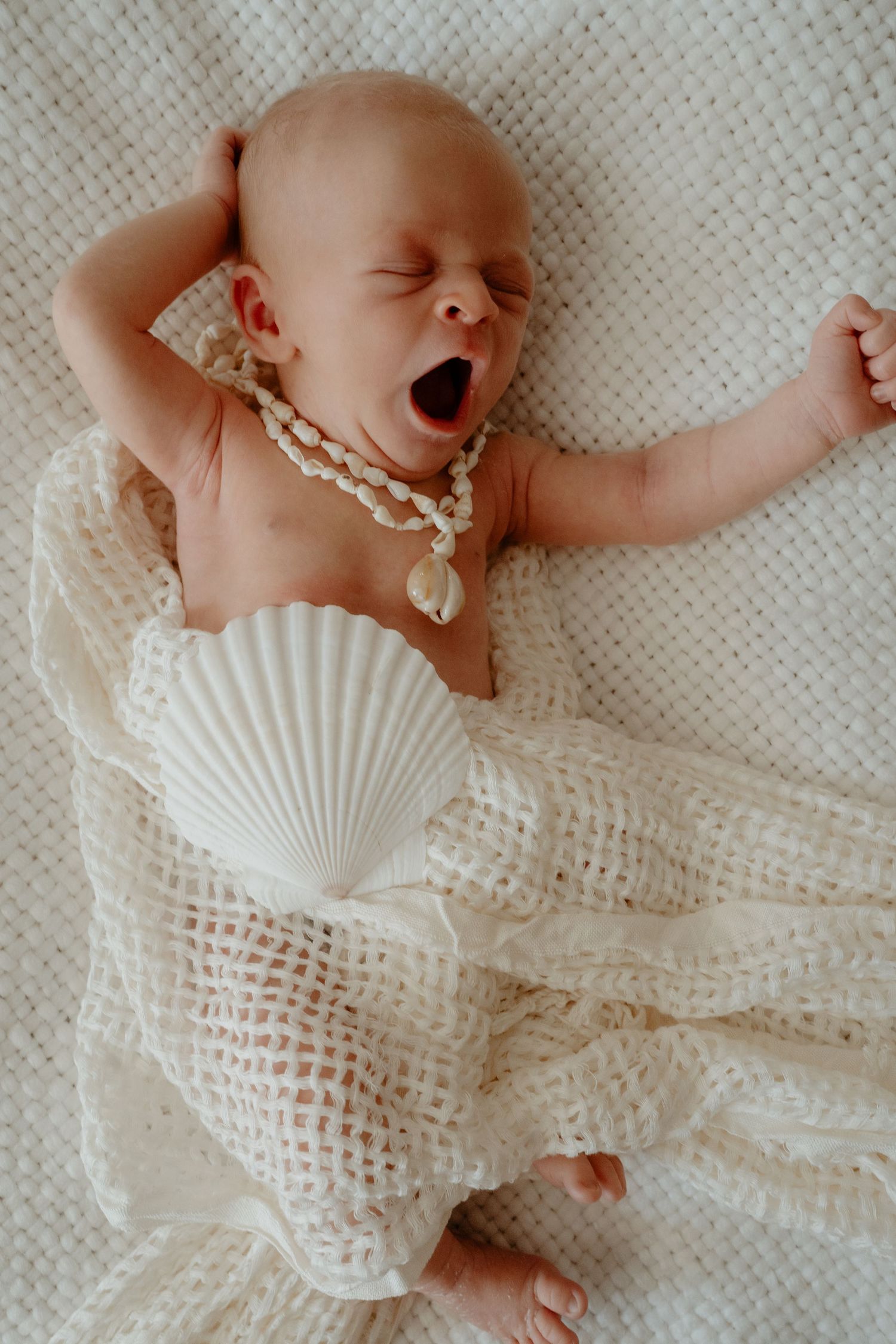 Yawning newborn baby adorned with seashell necklace lying on white knit blanket with large shell.