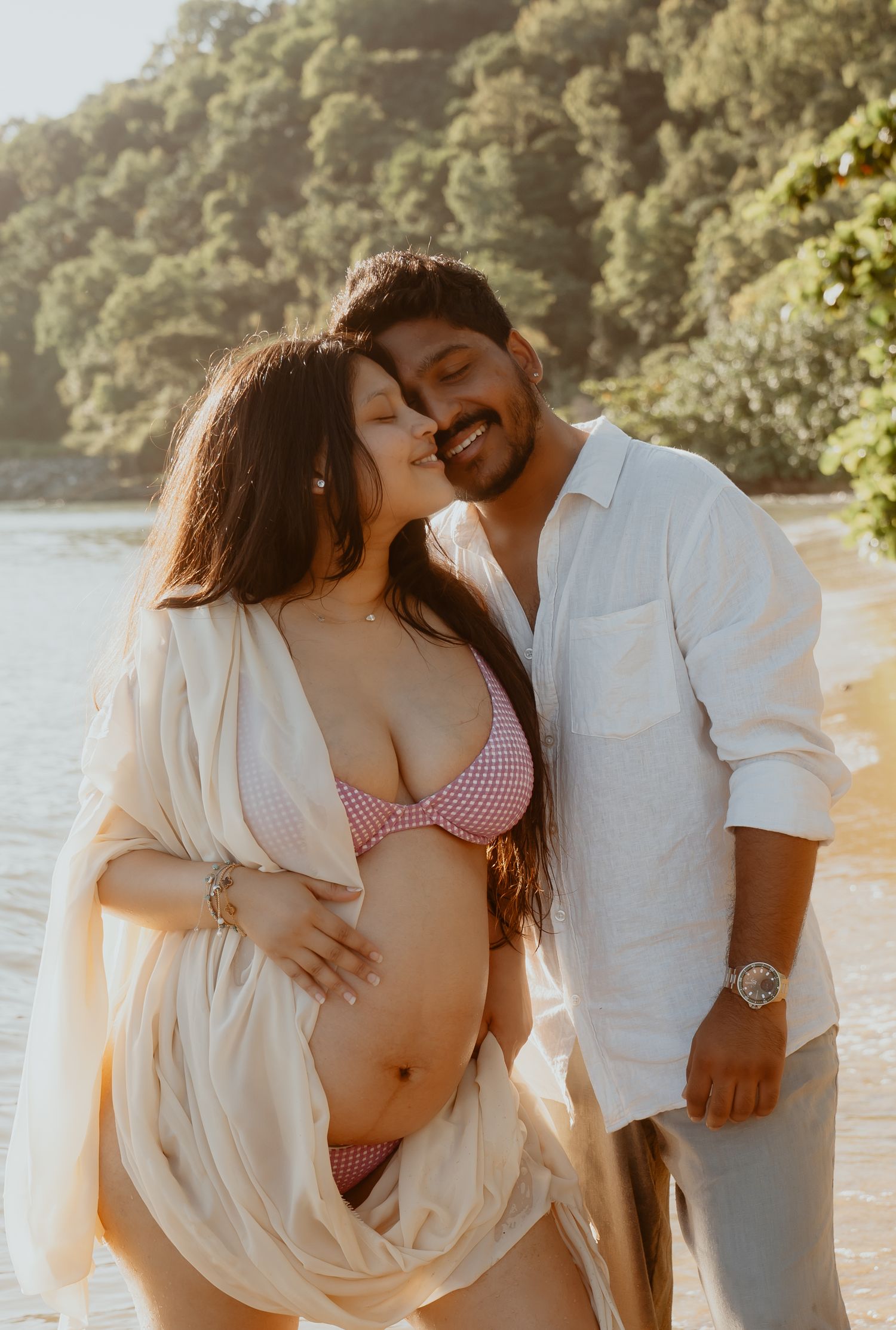 Smiling couple on a beach, pregnant woman in pink bikini embraced by man in white linen shirt.