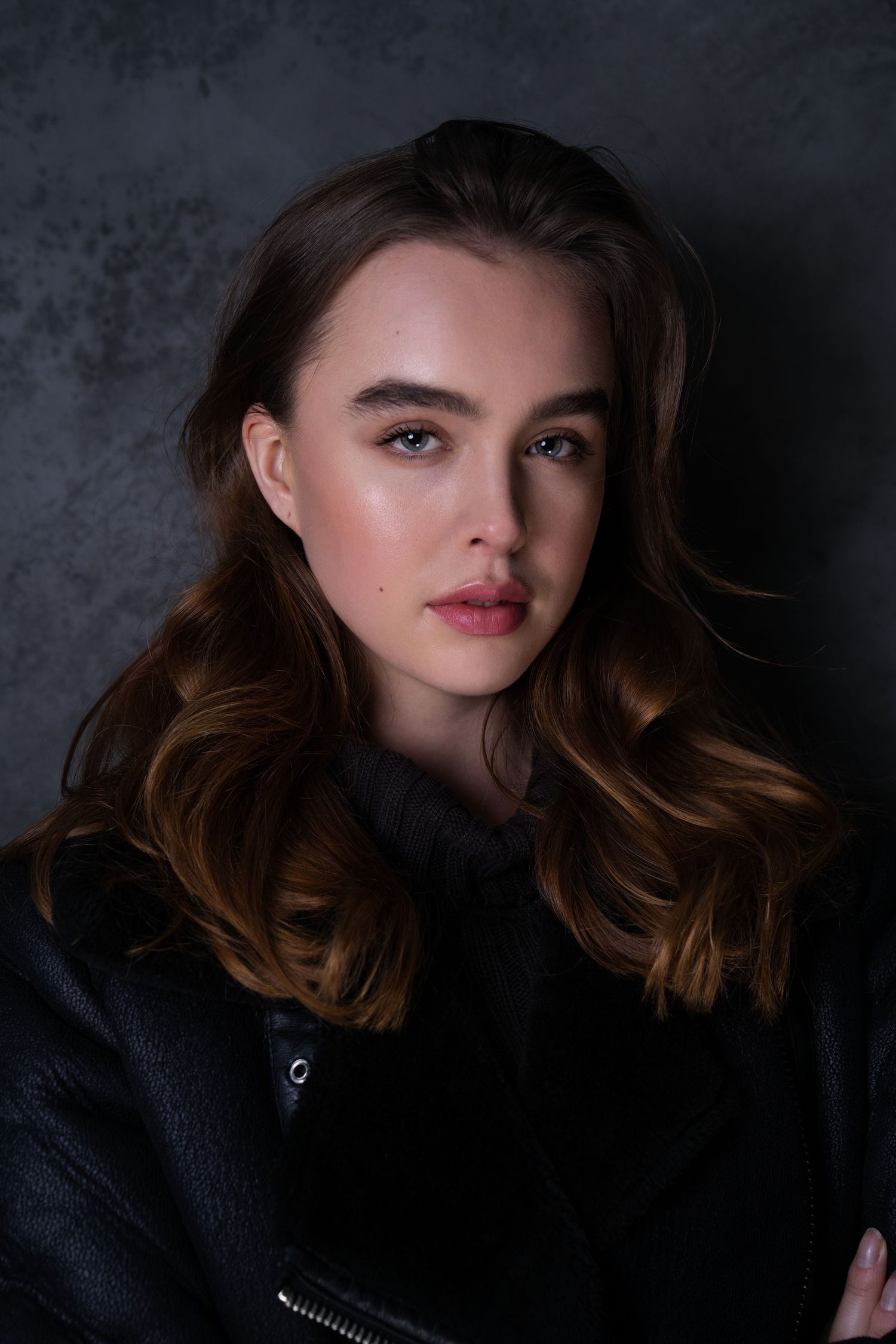 Close-up portrait of brunette woman in black leather jacket against dark gray textured studio background.