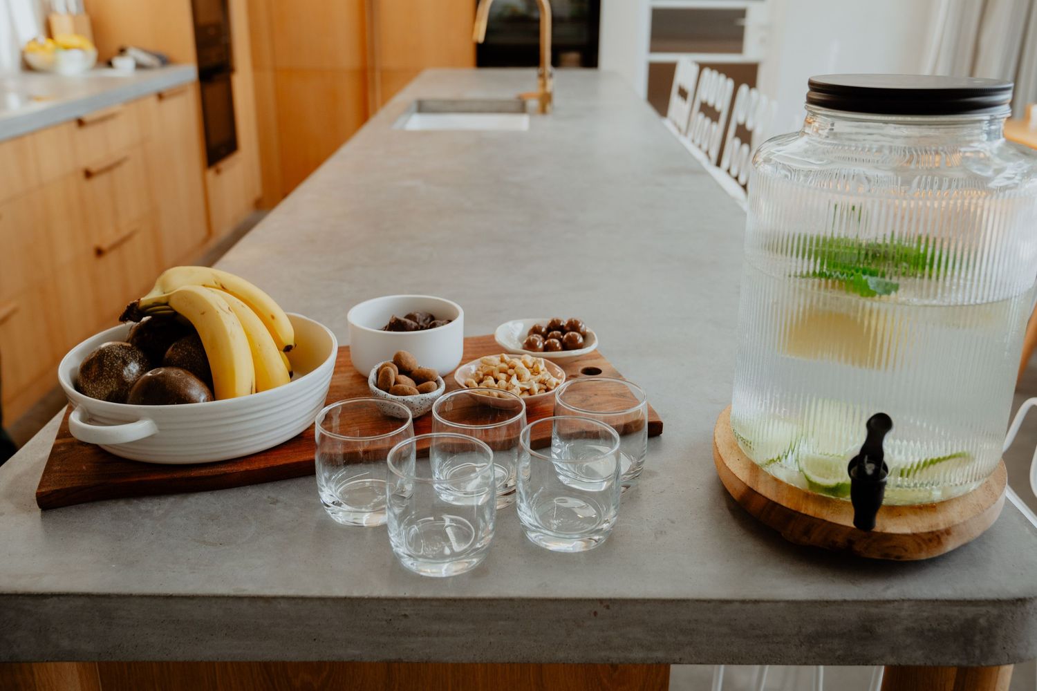 Healthy snack spread on a concrete countertop with fruits, nuts, glasses, and a large infused water dispenser.
