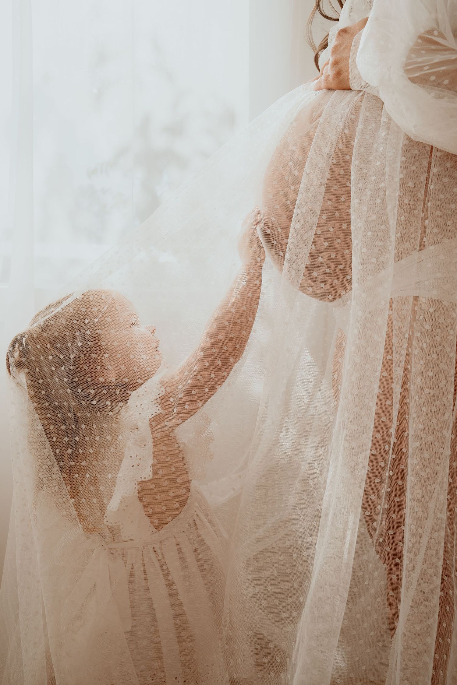 A young girl in a white dress gently touches a pregnant womans belly through a sheer polka dot maternity gown.