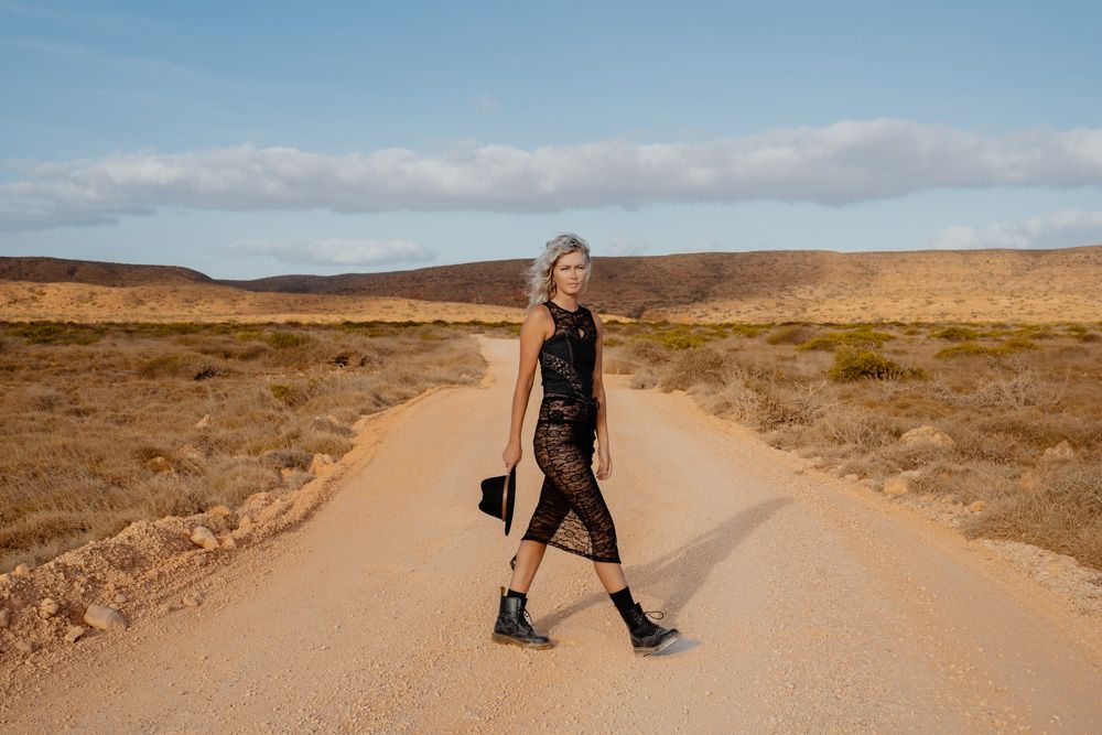 Woman in black dress and boots standing on a desert dirt road under a blue sky.