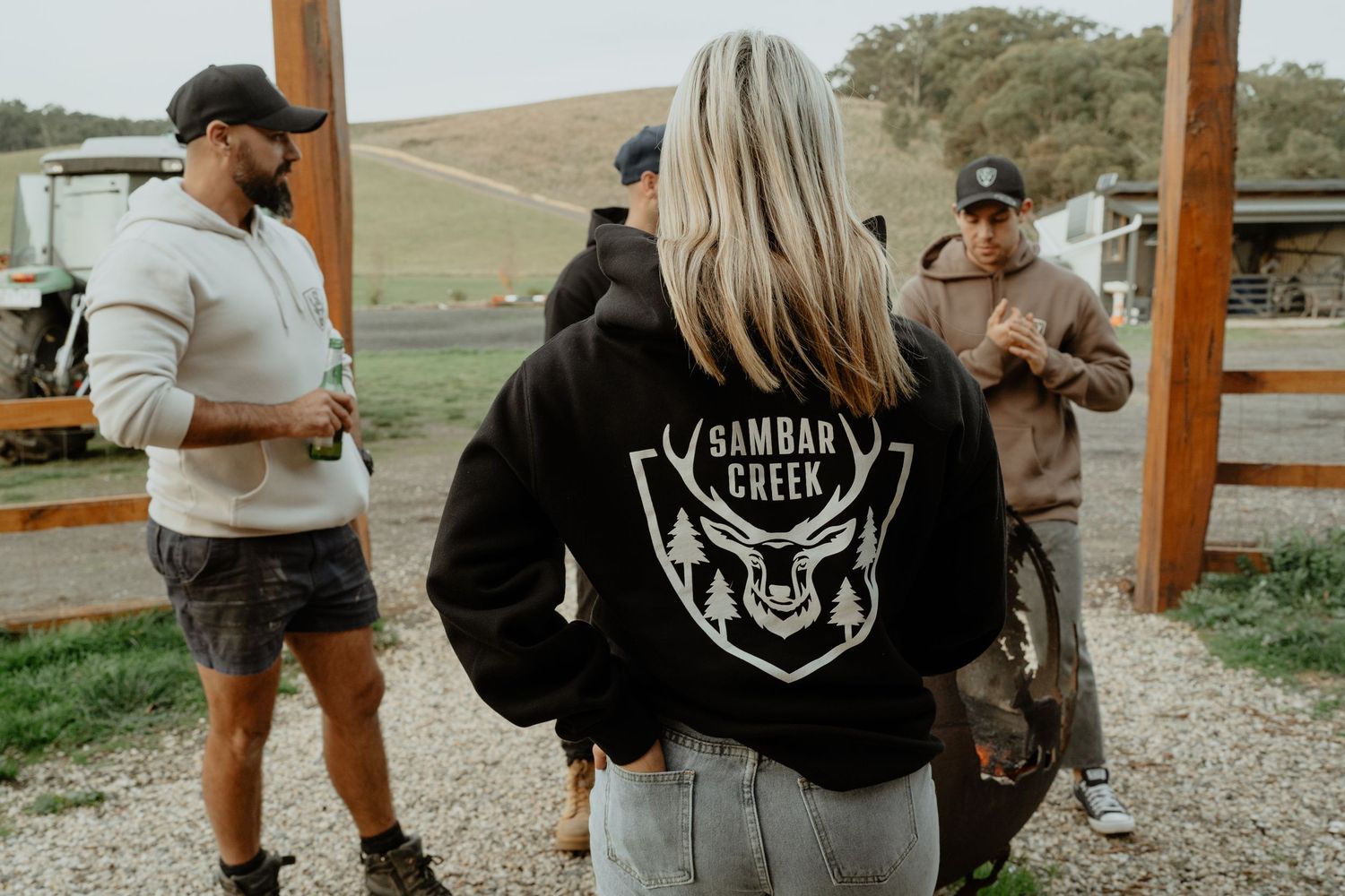 Group of friends wearing Sambar Creek hoodies socializing outdoors at a rural farm setting.