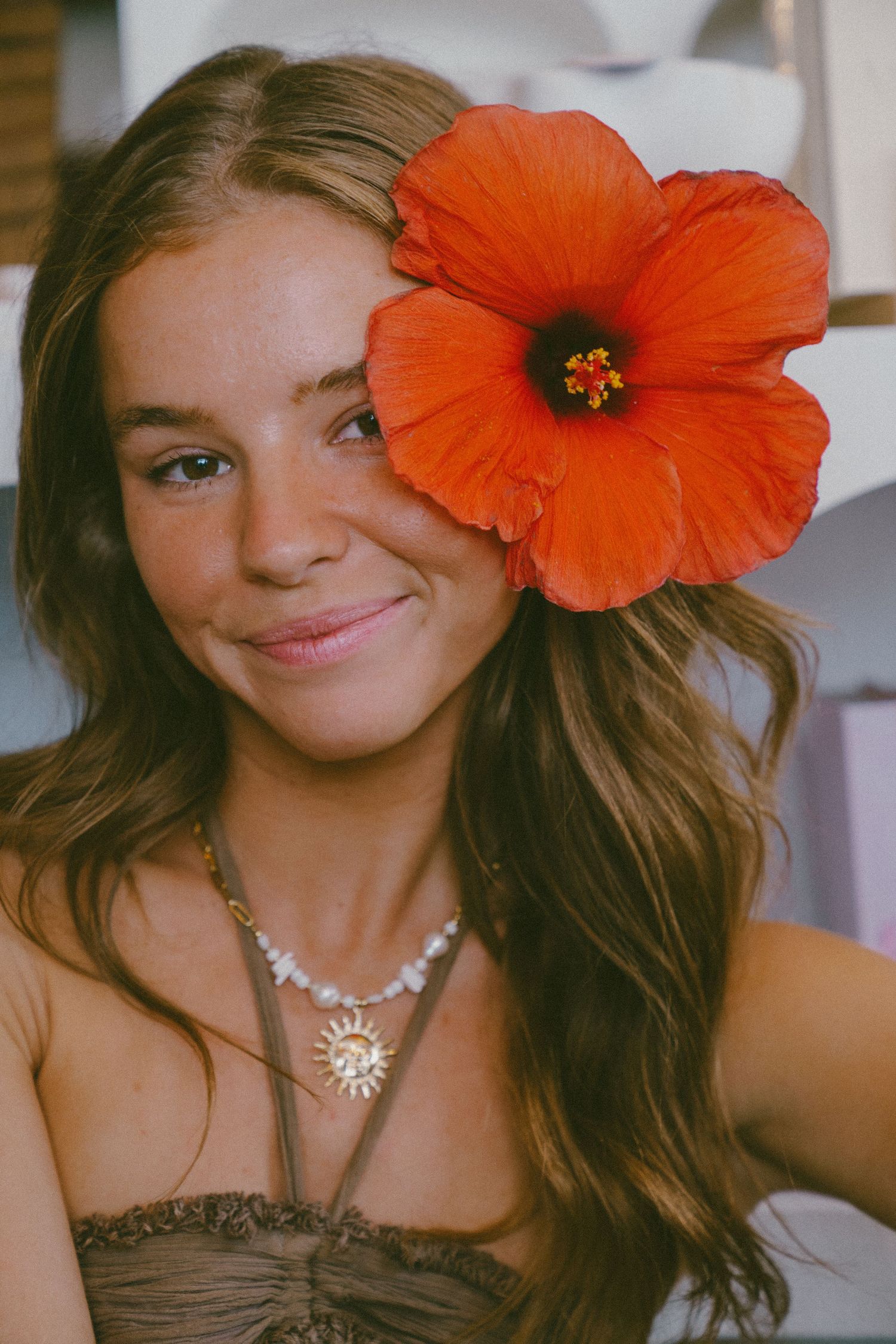 Young woman smiling with a large red hibiscus flower in her hair and a sun pendant necklace.
