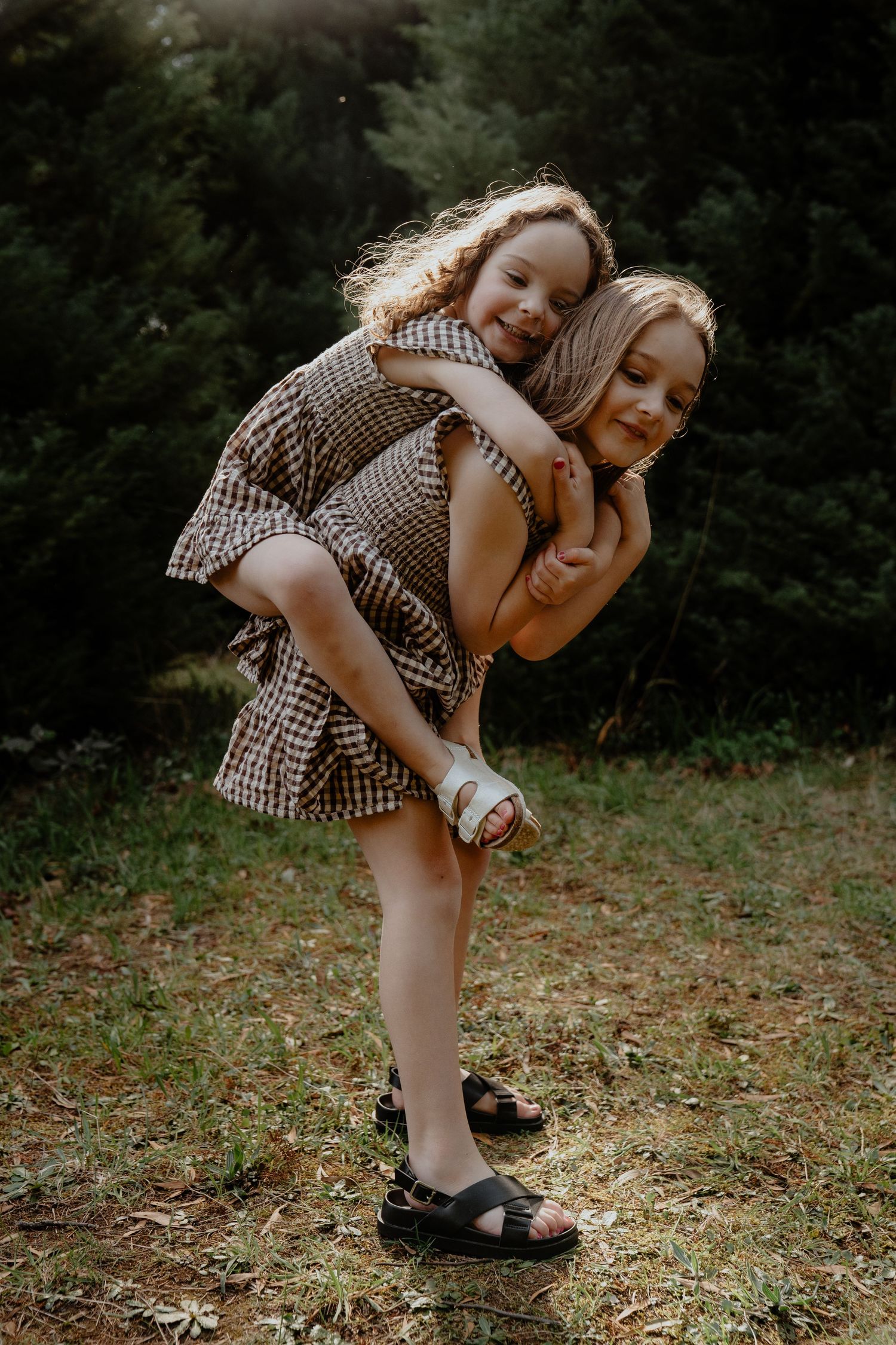 Two sisters in matching gingham dresses share a piggyback hug in forest clearing.