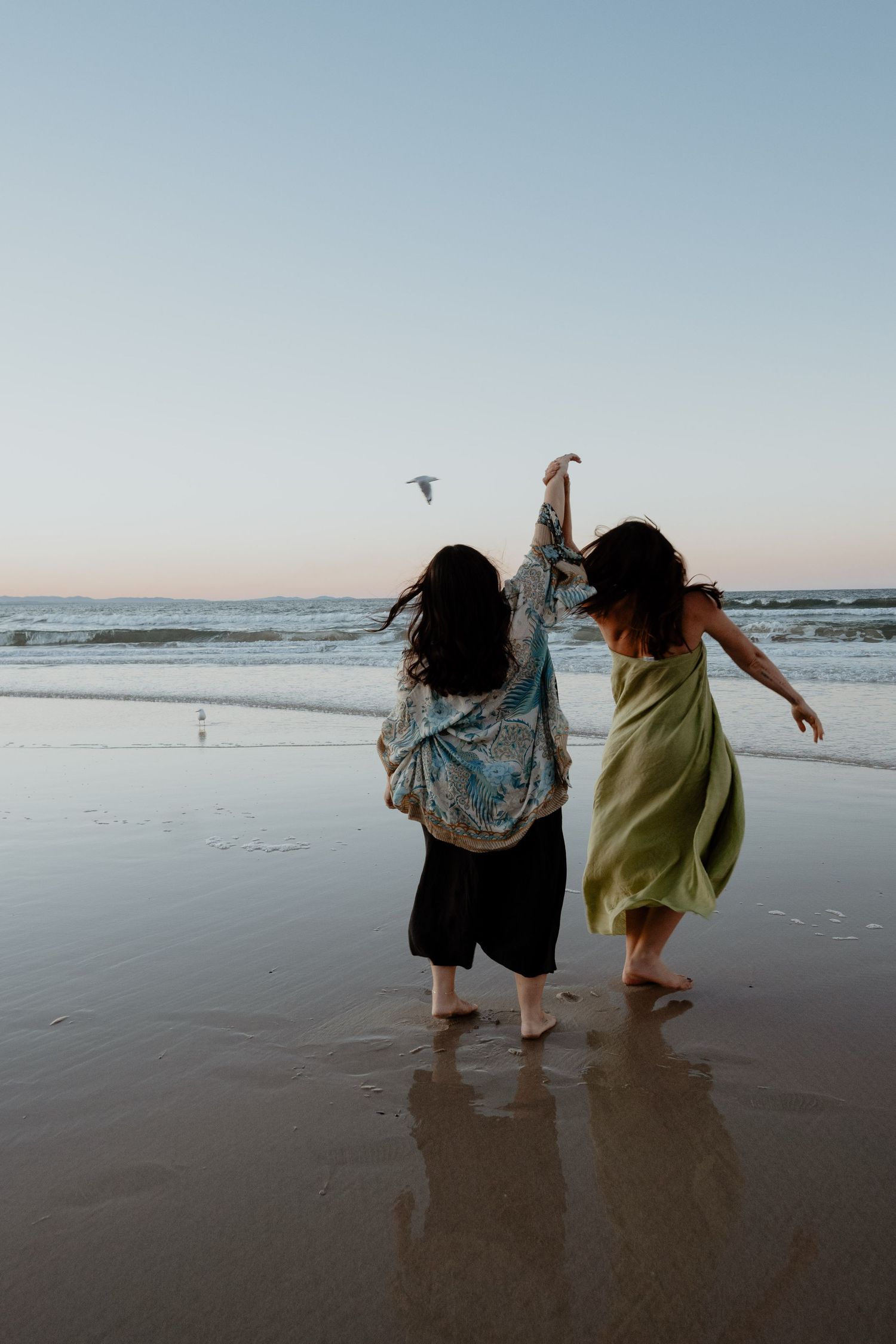 Two women dancing barefoot on a wet beach at dusk, facing the ocean with arms raised.