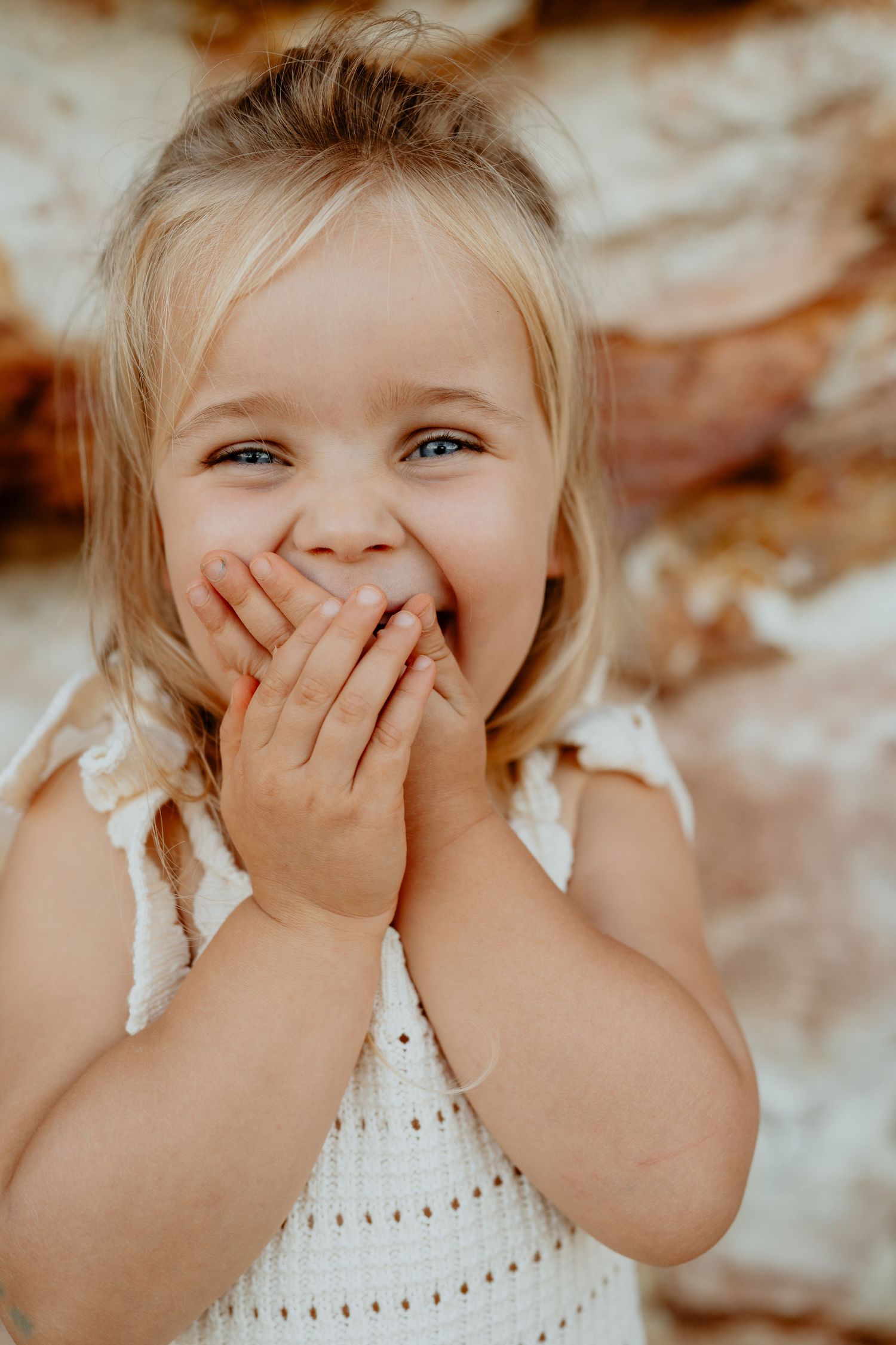 Playful blonde toddler girl covering her mouth with both hands and giggling against a rocky background.