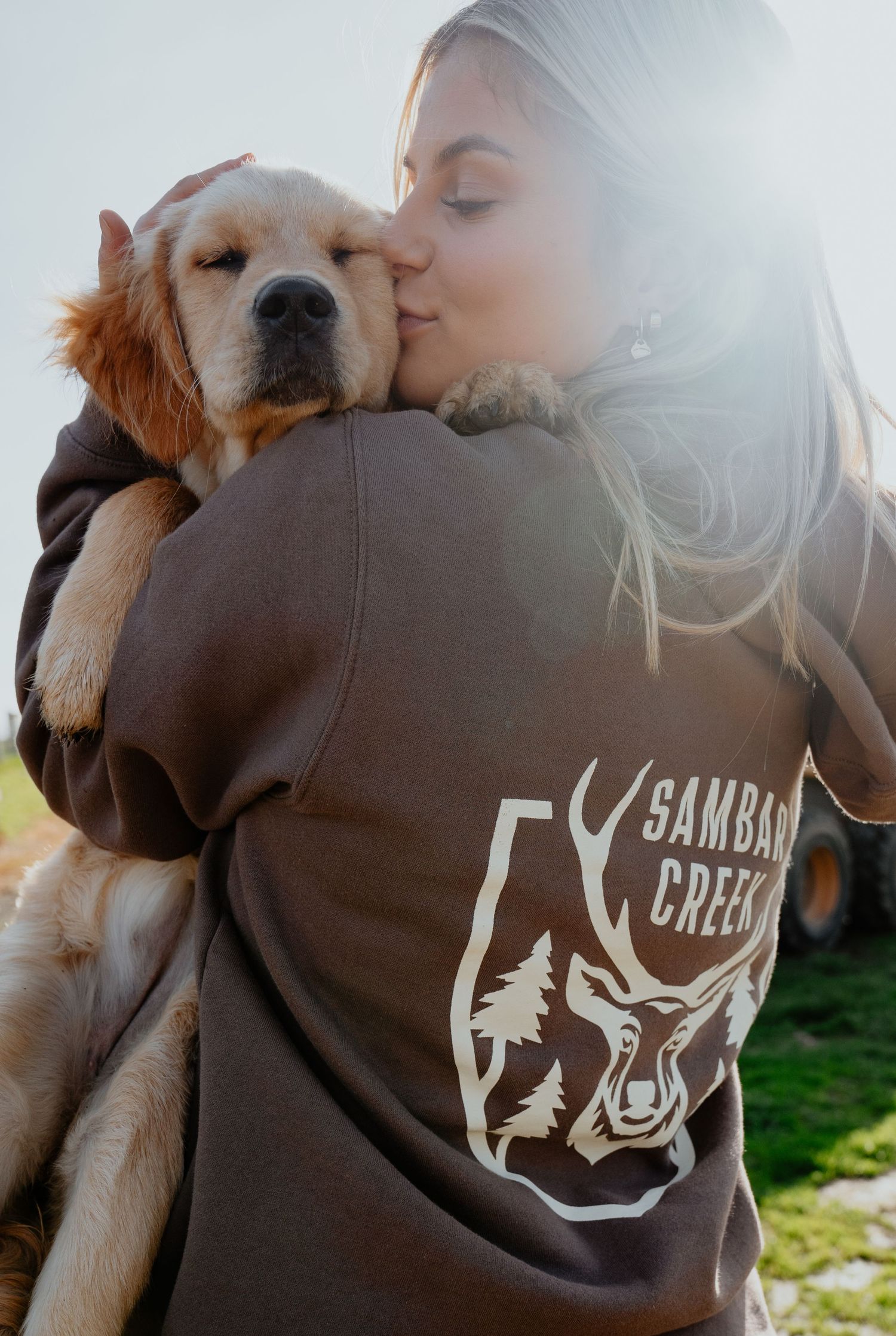 Woman in Sambar Creek brown hoodie cuddling a golden retriever puppy in sunny outdoor setting.