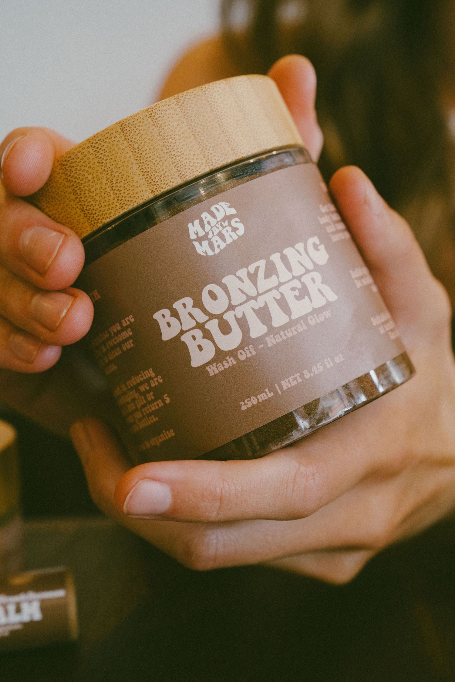 Hands holding a jar of Made by Mars Bronzing Butter natural self-tanner with bamboo lid, 250ml.