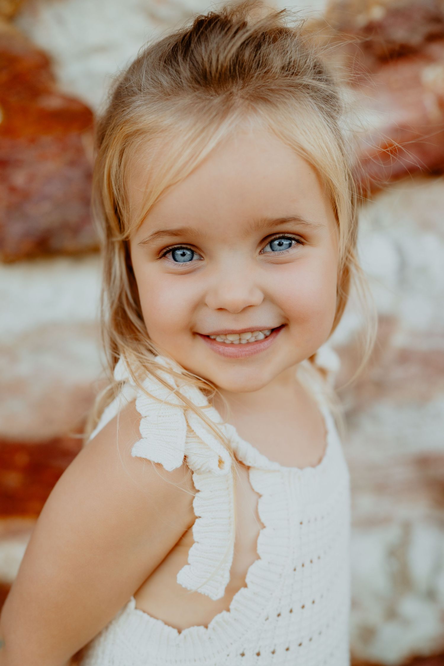 Cute blonde blue-eyed little girl in white crochet dress smiling sweetly against a rocky background.