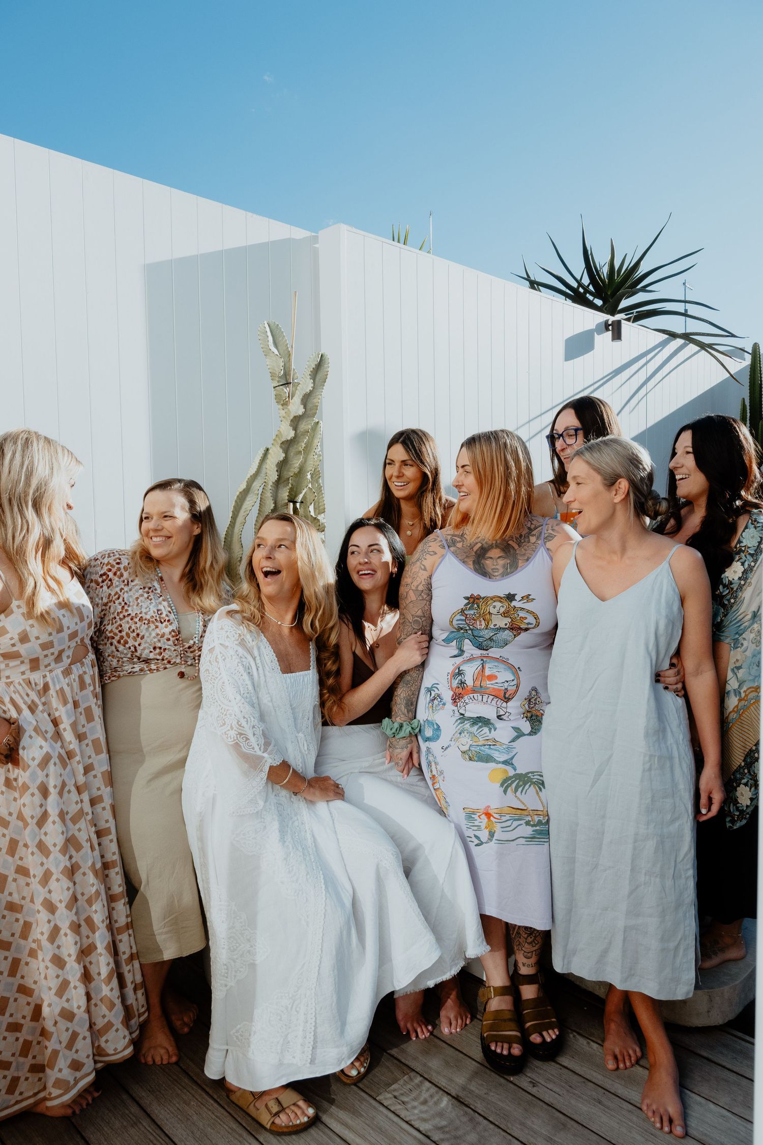 Group of women smiling and laughing together on a sunny rooftop with cactus decor and blue sky.