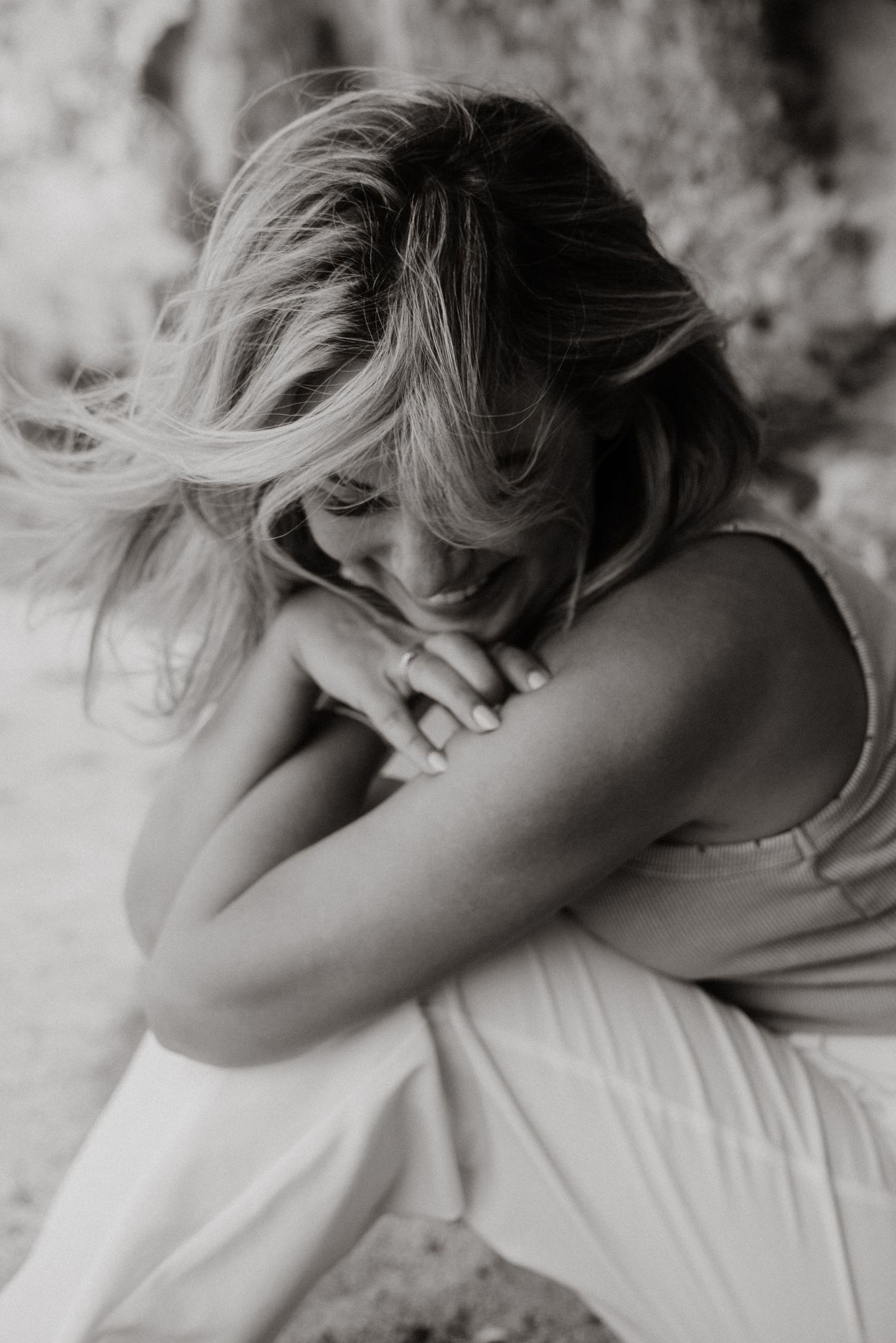 Black and white photo of a blonde woman hugging her knees near a rocky backdrop.