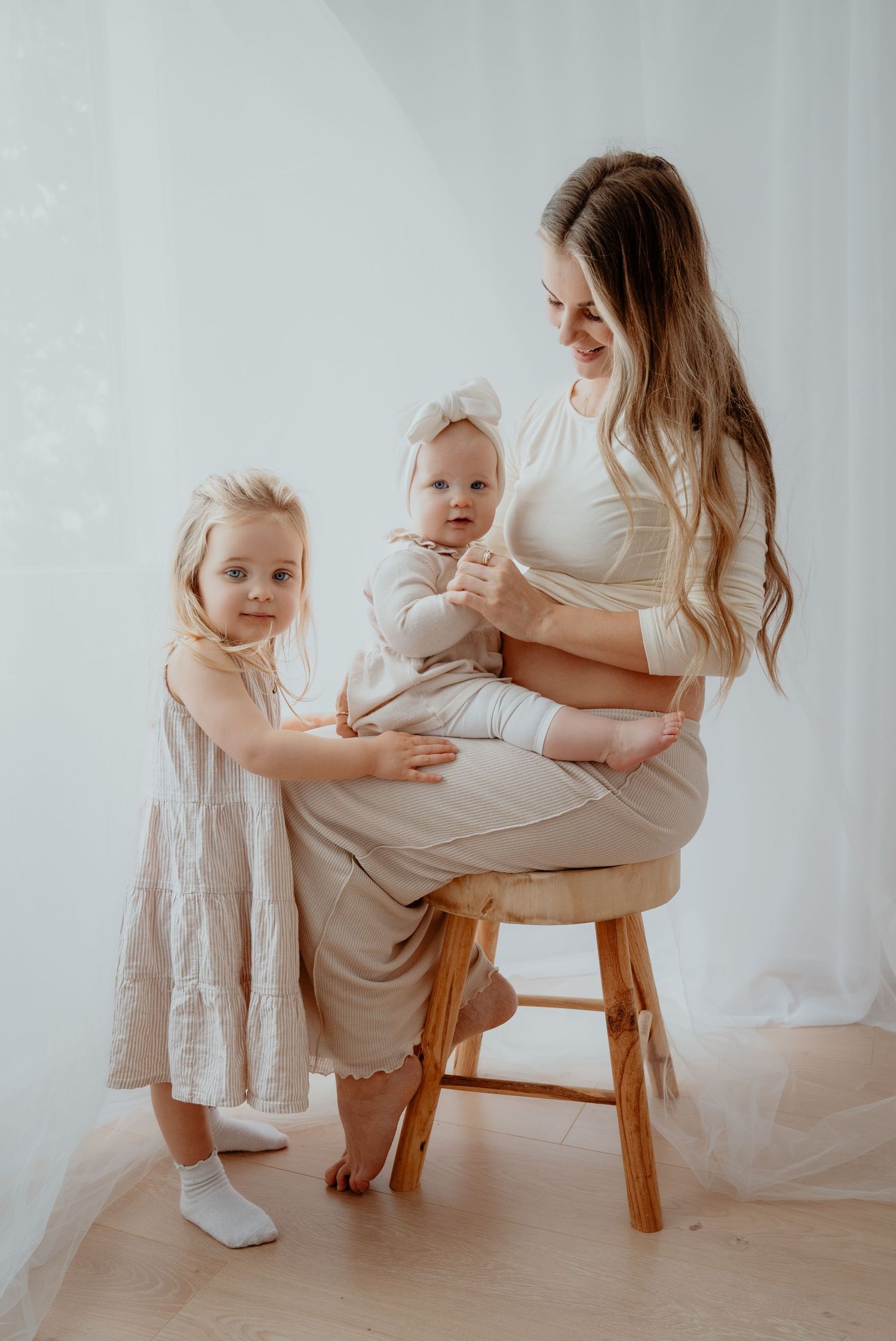 A mother with long blonde hair sits on a wooden stool holding a baby while a young girl stands beside her smiling.