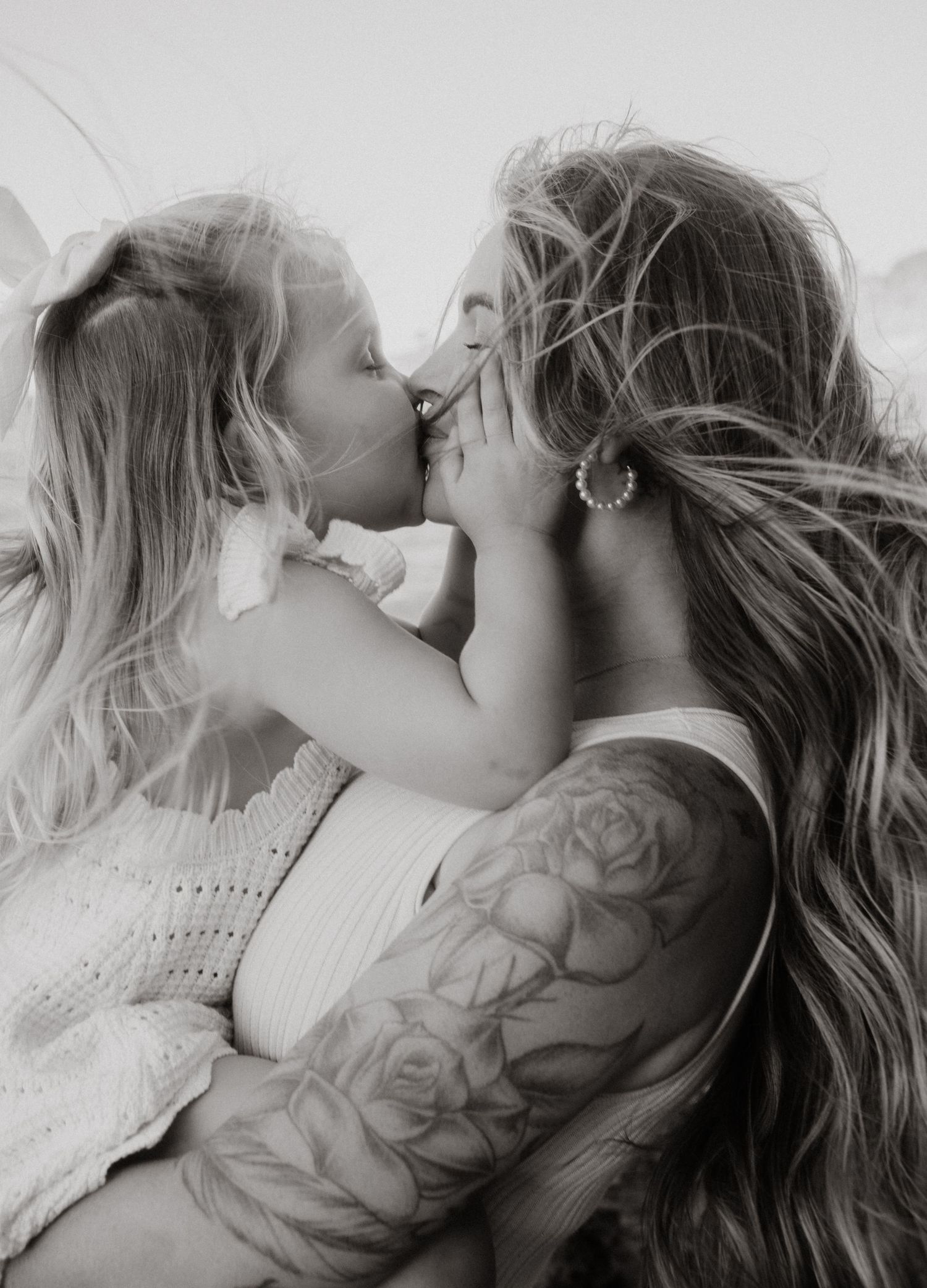Black and white photo of tattooed mother receiving a kiss from her young daughter.