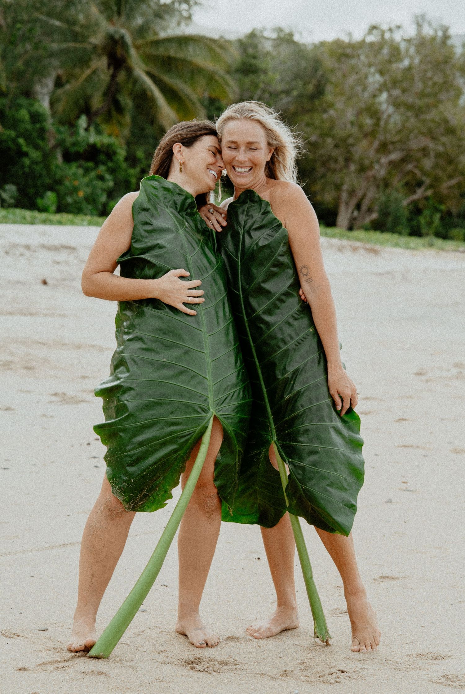 Two women laughing and hugging while holding large tropical green leaves on a sandy beach.