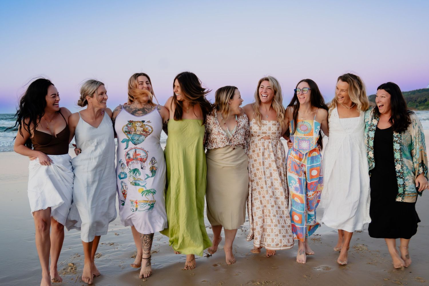 Group of nine women in colorful summer dresses laughing together on a beach at sunset.