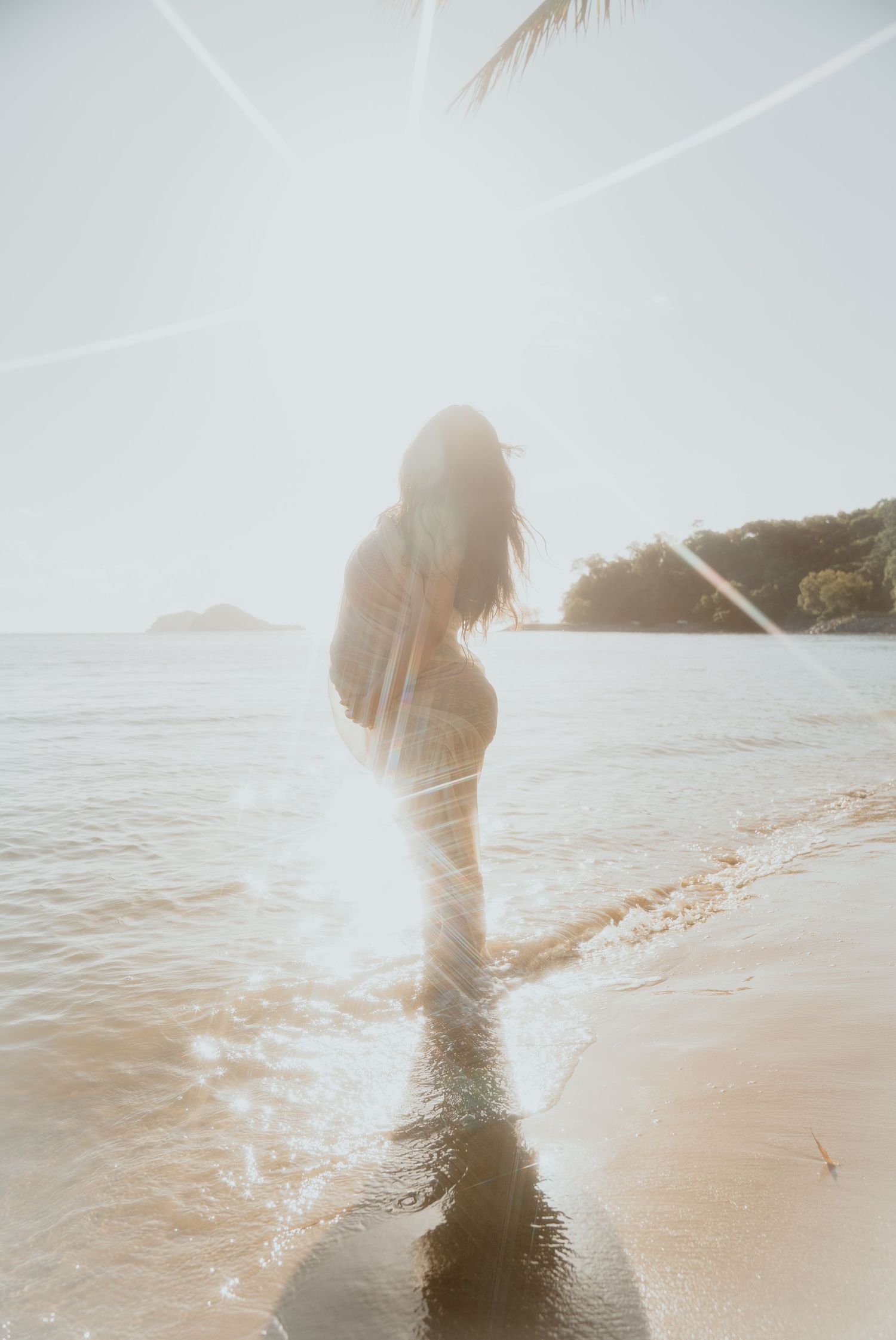 Pregnant woman standing in shallow ocean waves on a bright sunlit tropical beach with lens flare.