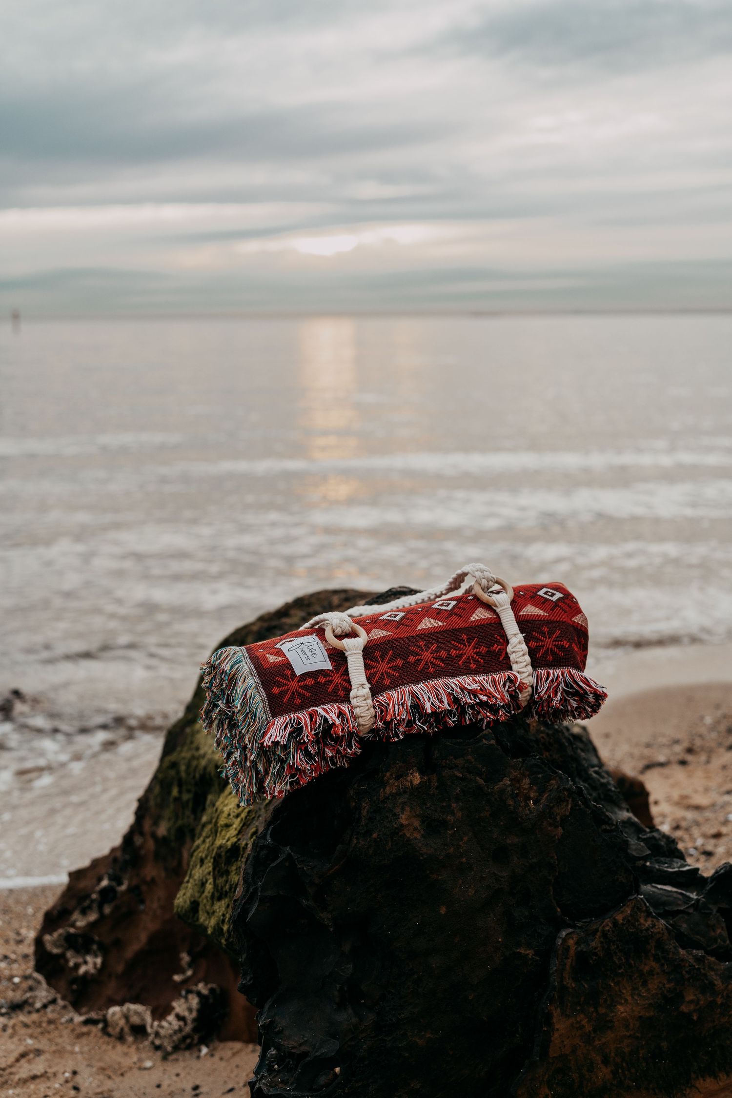 Red geometric patterned fringed duffle bag resting on mossy rock at overcast beach.