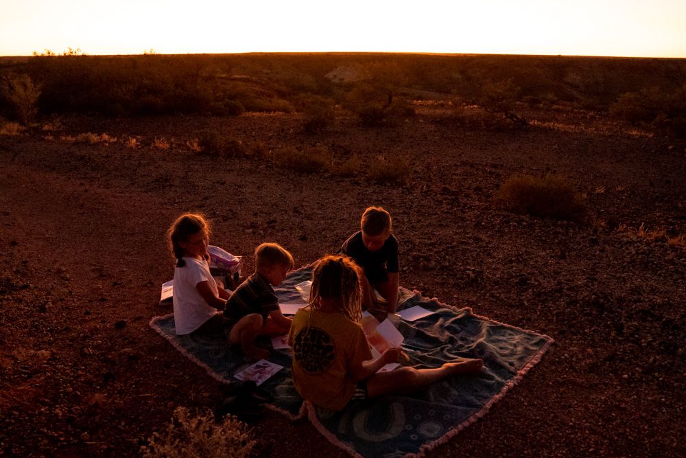 Children sitting on a blanket watching a stunning golden sunset over a red desert landscape.