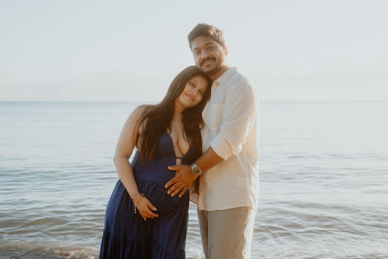 Couple embracing on beach at sunset, pregnant woman in blue dress with partner holding her belly.