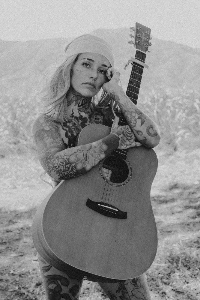 Tattooed blonde woman holding an acoustic guitar in a moody black and white desert portrait.