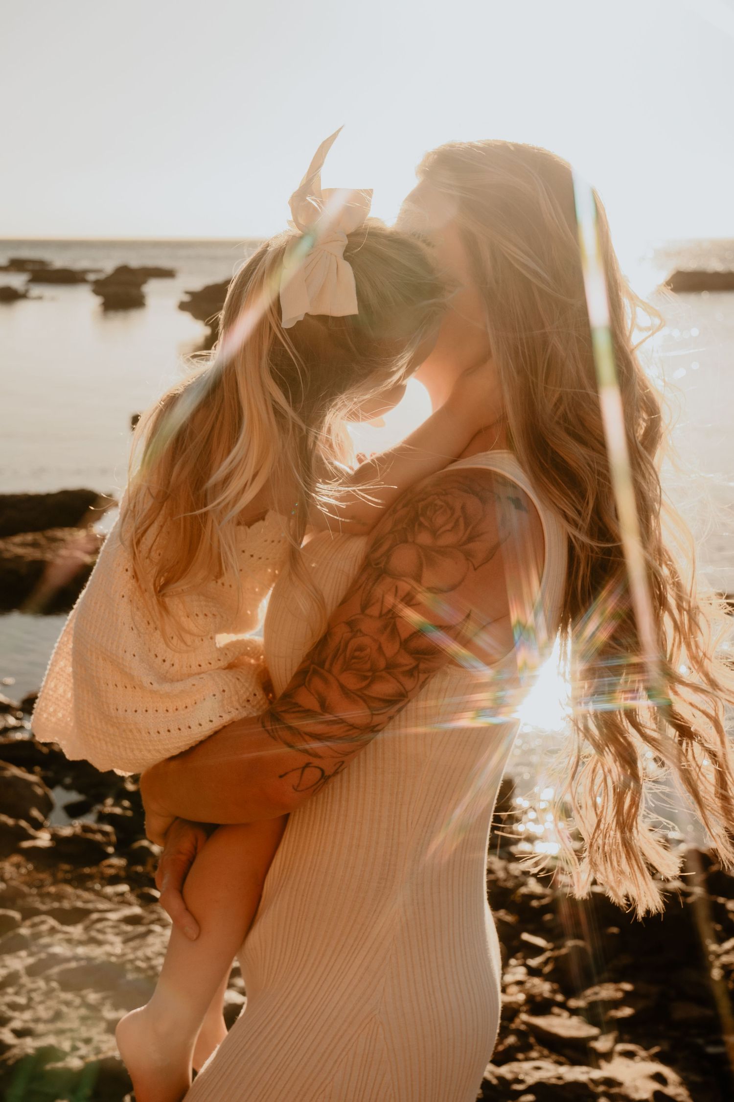 Mother hugging her young daughter with a bow in her hair on a rocky beach with sun flare.