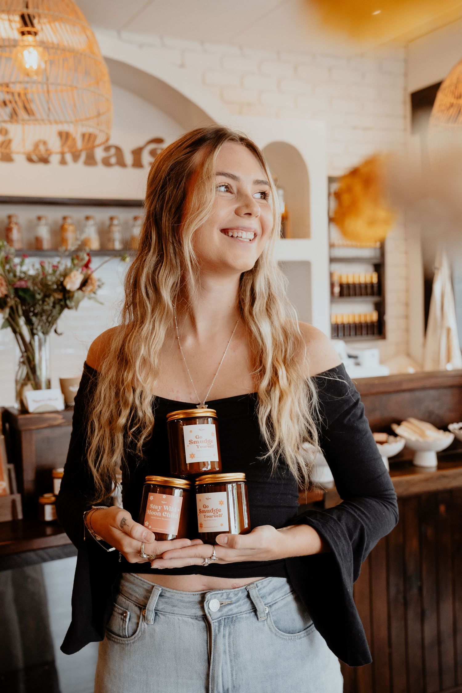 Smiling blonde woman holding three stacked glass jars of product inside Remi and Mars boutique.