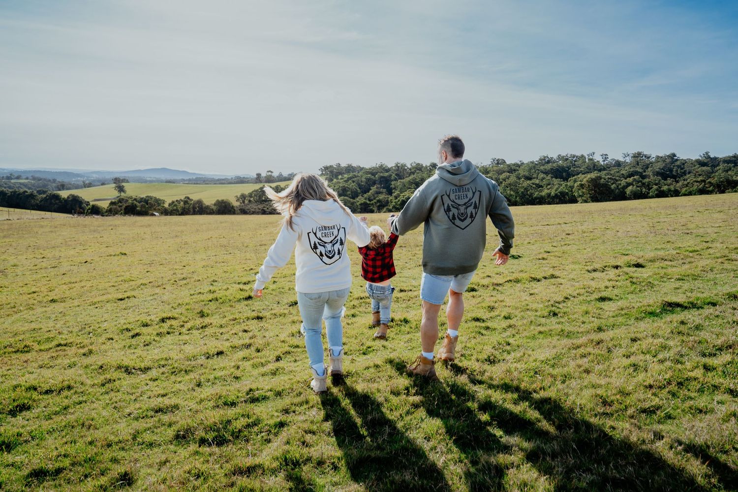 Family of three walking hand in hand across a green open field on a sunny day.