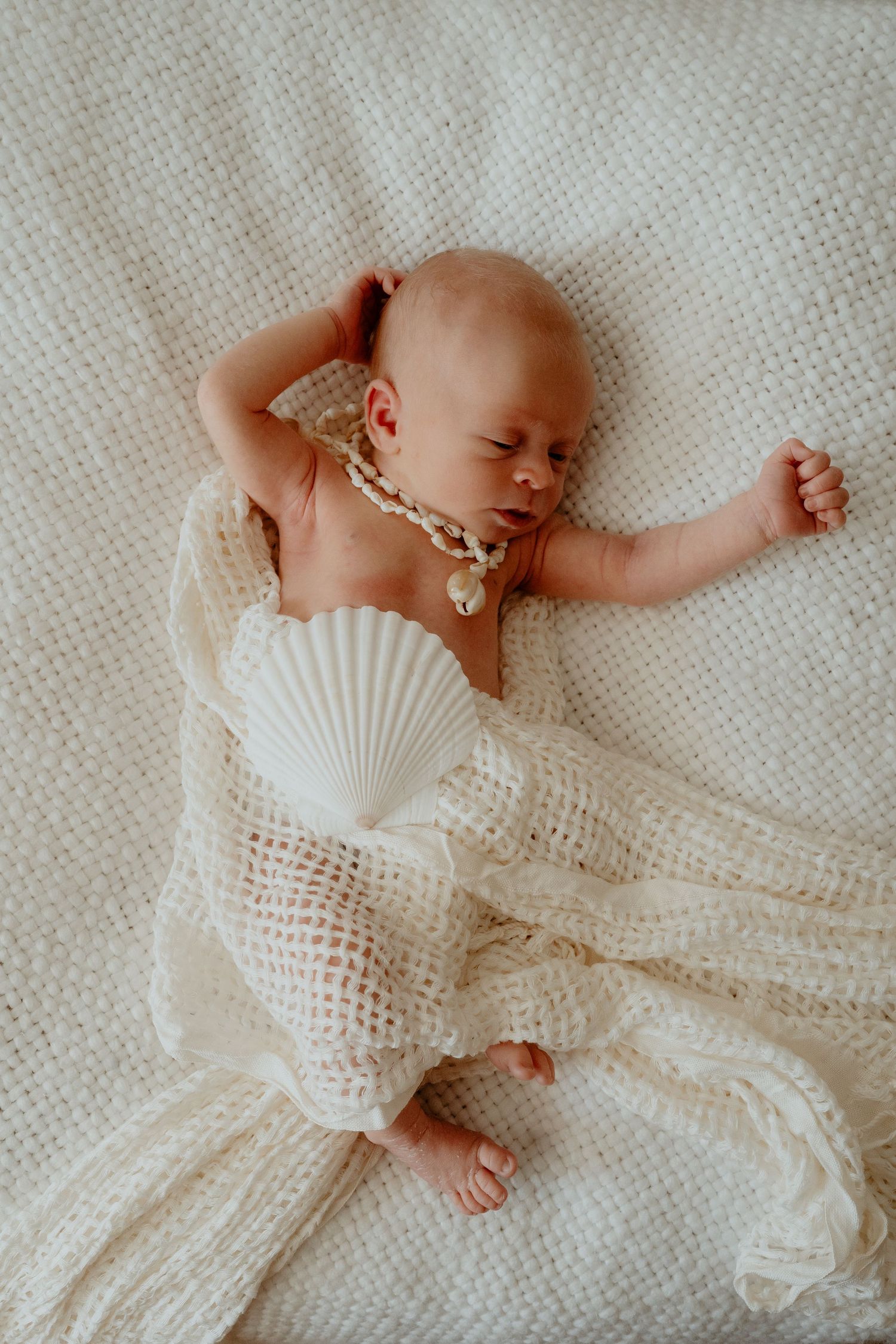 Sleeping newborn baby wrapped in cream crochet blanket with seashell and pearl necklace on white knit background.