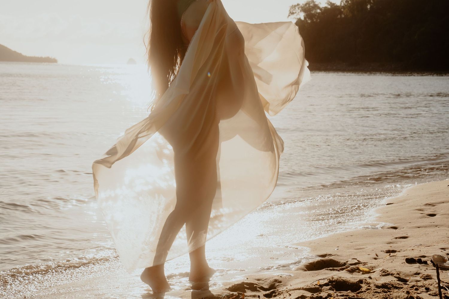 Pregnant woman walking on sunny beach holding flowing sheer white fabric in golden sunlight.