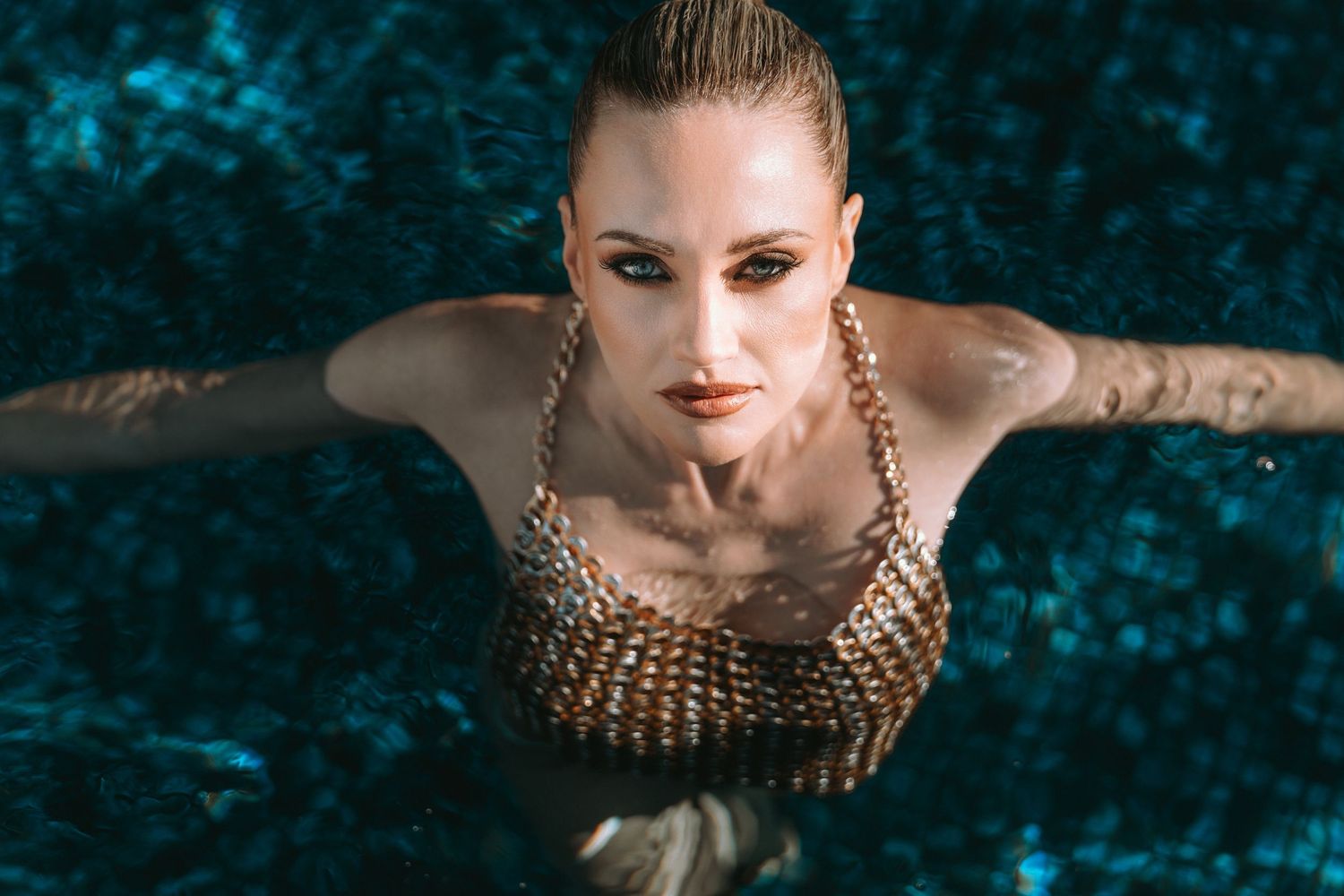 Aerial view of a wet woman in a gold chainmail swimsuit emerging from a dark blue pool.