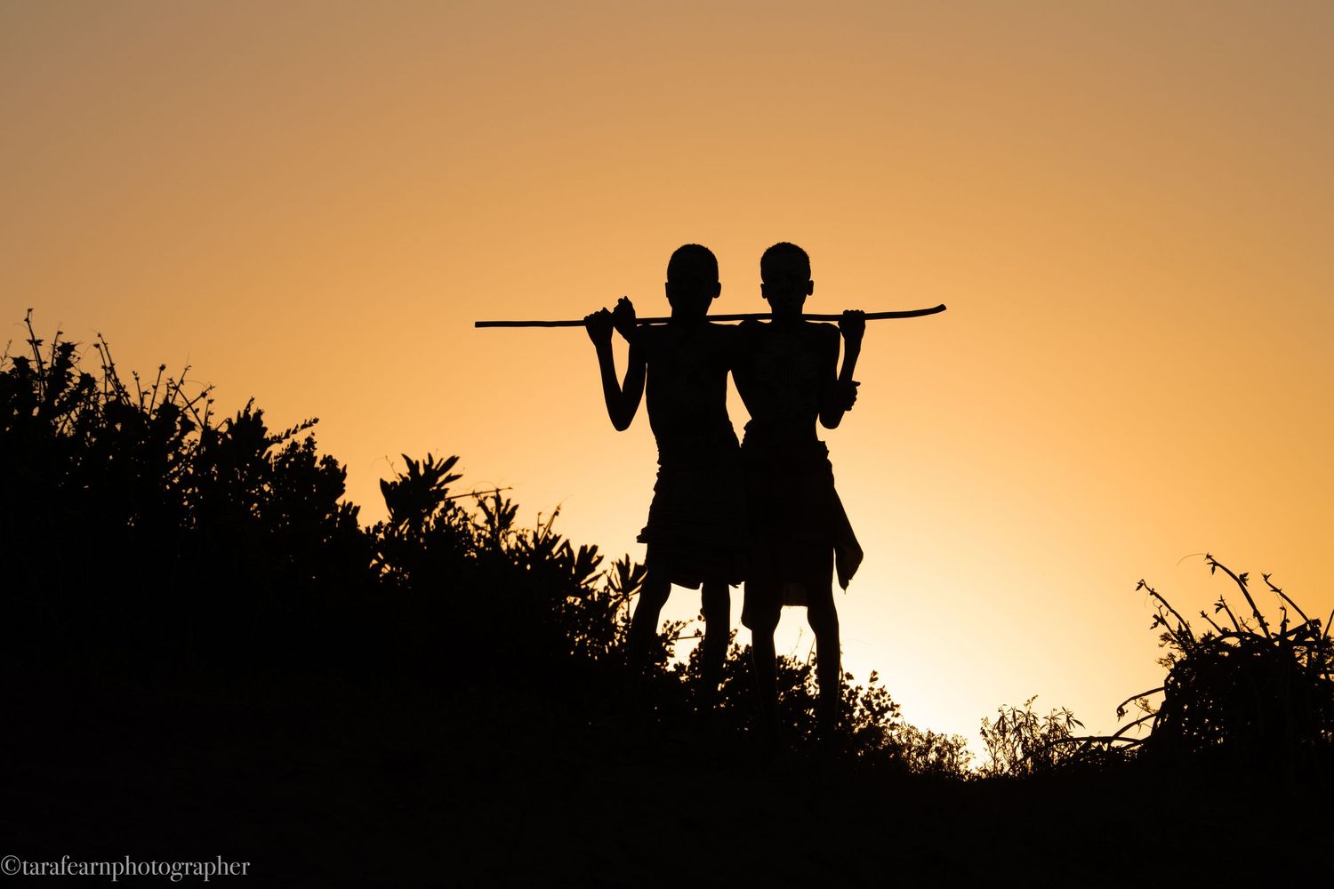 Silhouette of two people carrying a long pole together against a vibrant golden sunset sky.