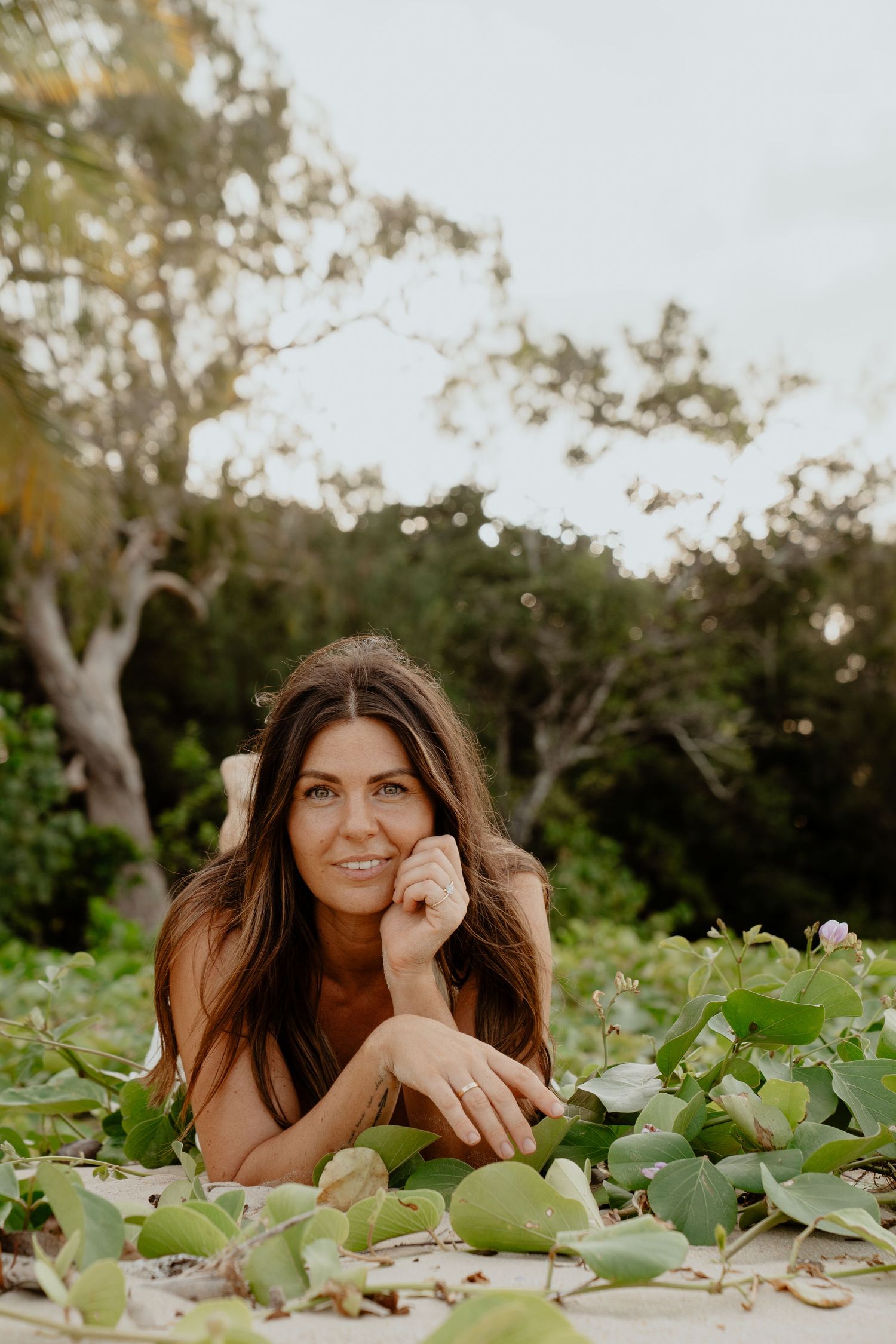 Brunette woman lying among green beach vines smiling at camera in natural setting.