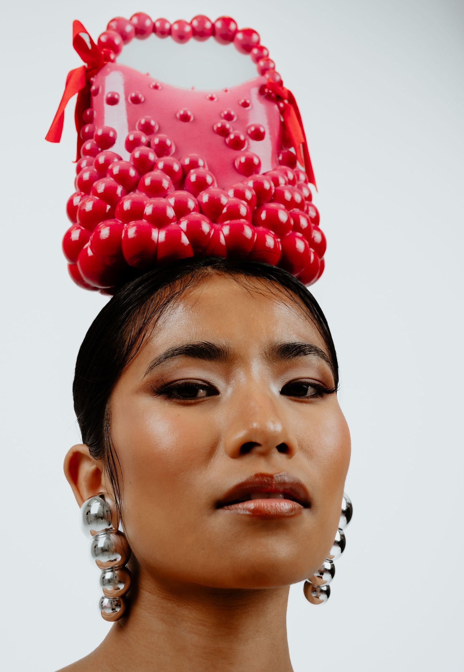 Model wearing red beaded handbag on head with silver earrings, fashion editorial style.