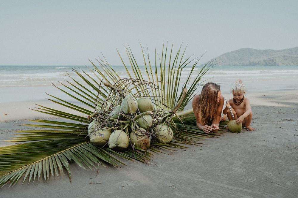 Mother and child crouching together examining fresh coconuts on a tropical sandy beach.