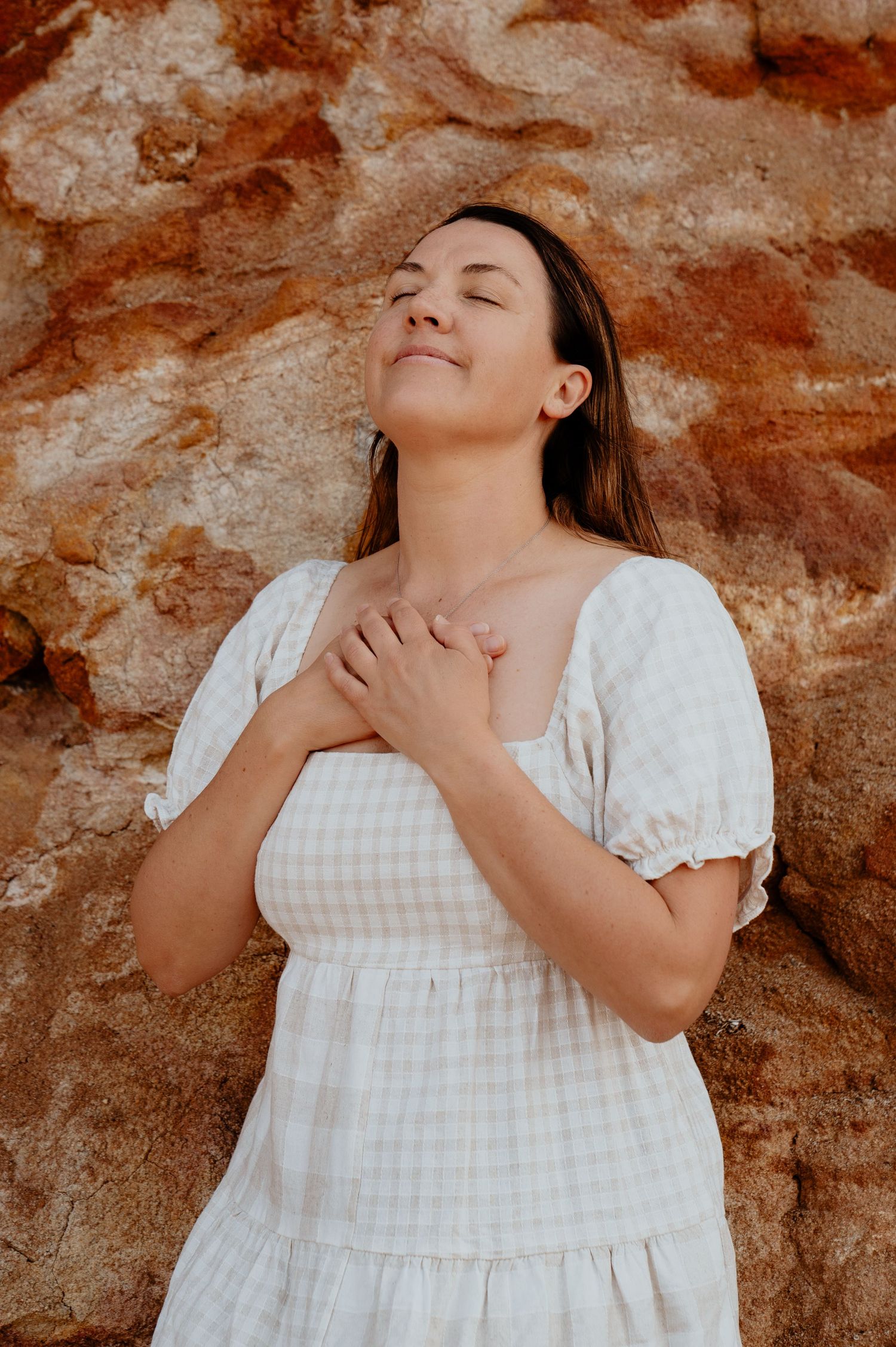 Woman in white gingham dress leans against red rock wall with eyes closed, hands on chest.