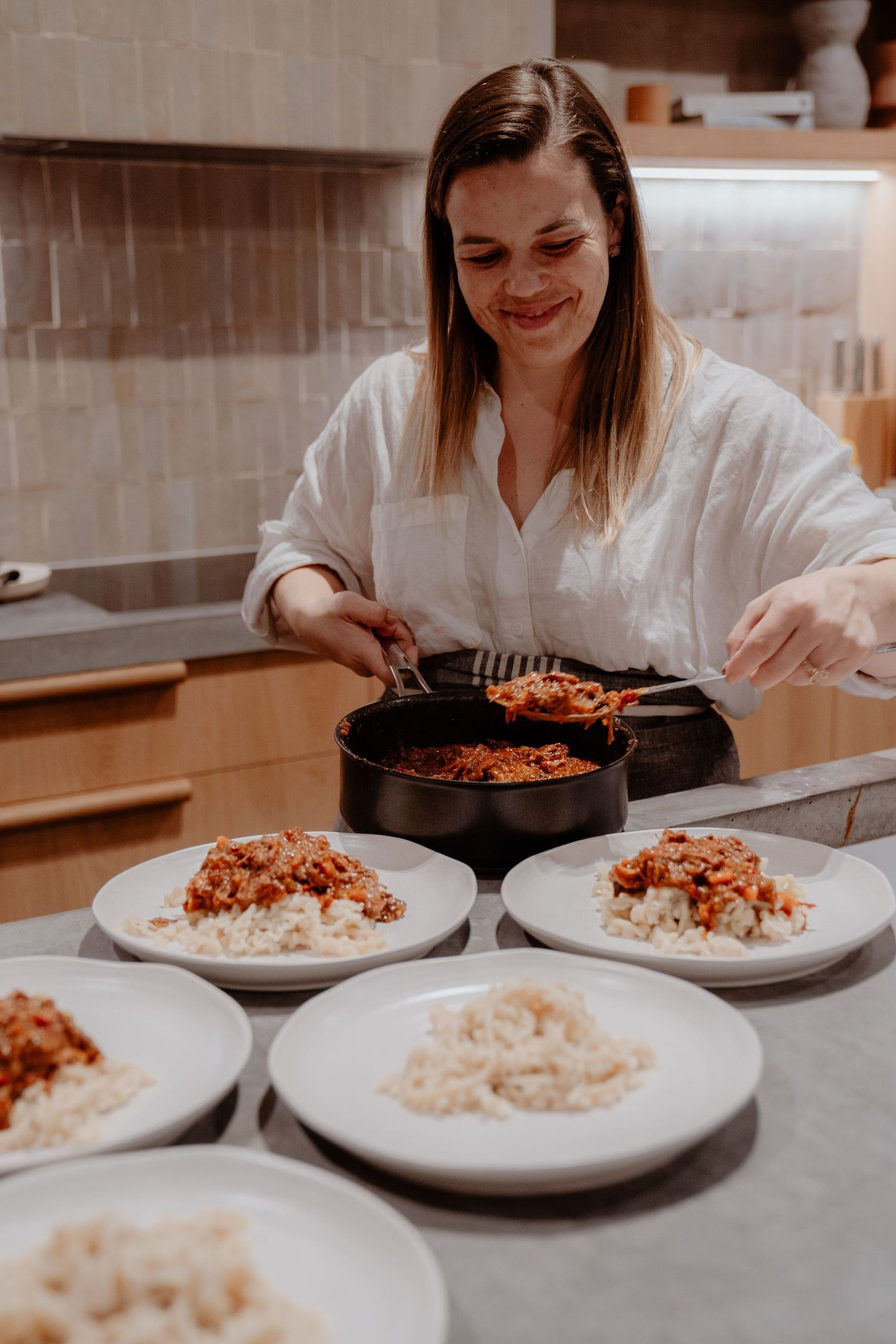 Smiling woman in white shirt serving meat sauce over rice on multiple white plates in a modern kitchen.