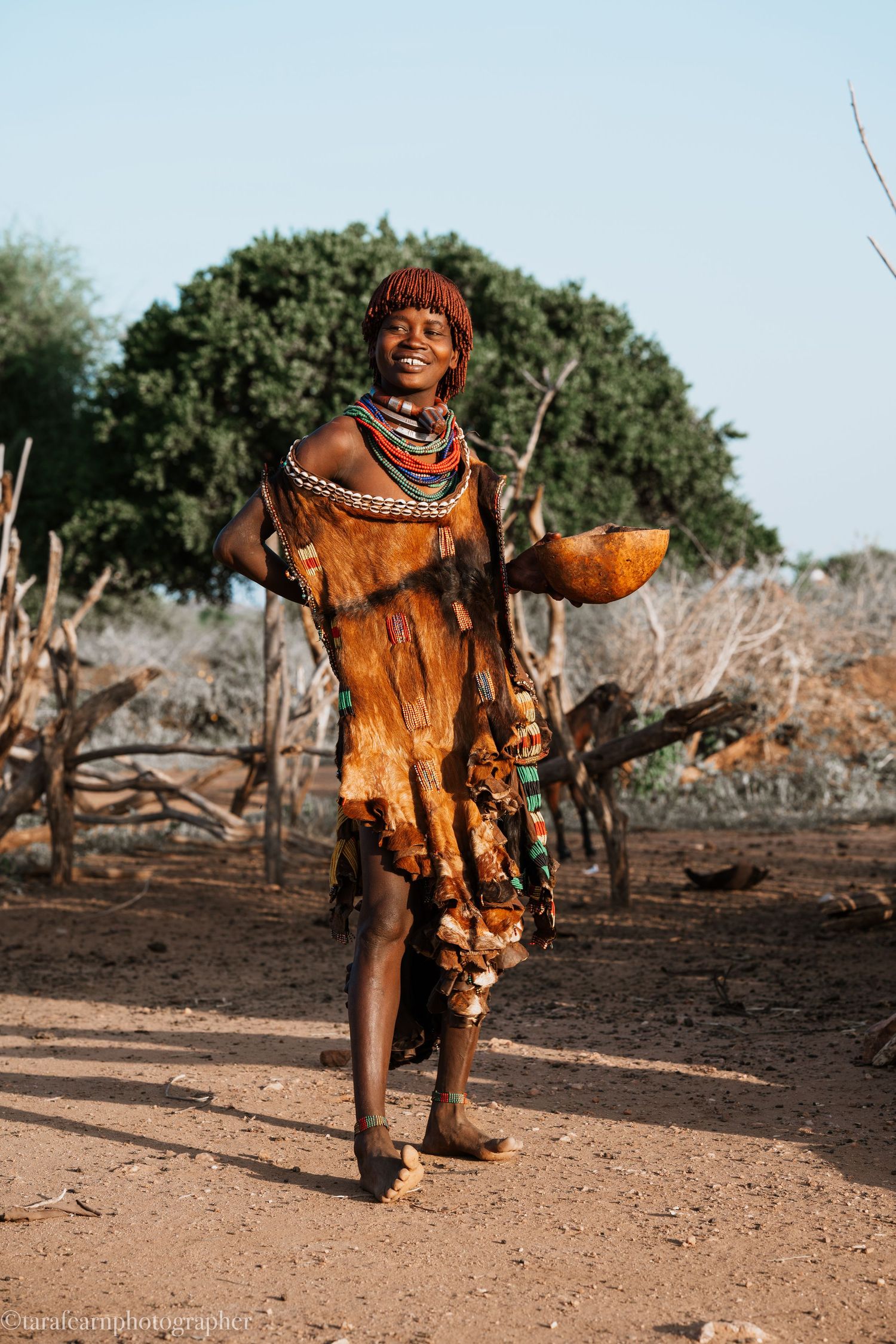 Smiling African tribal woman in traditional leather dress and colorful beaded necklaces standing outdoors.