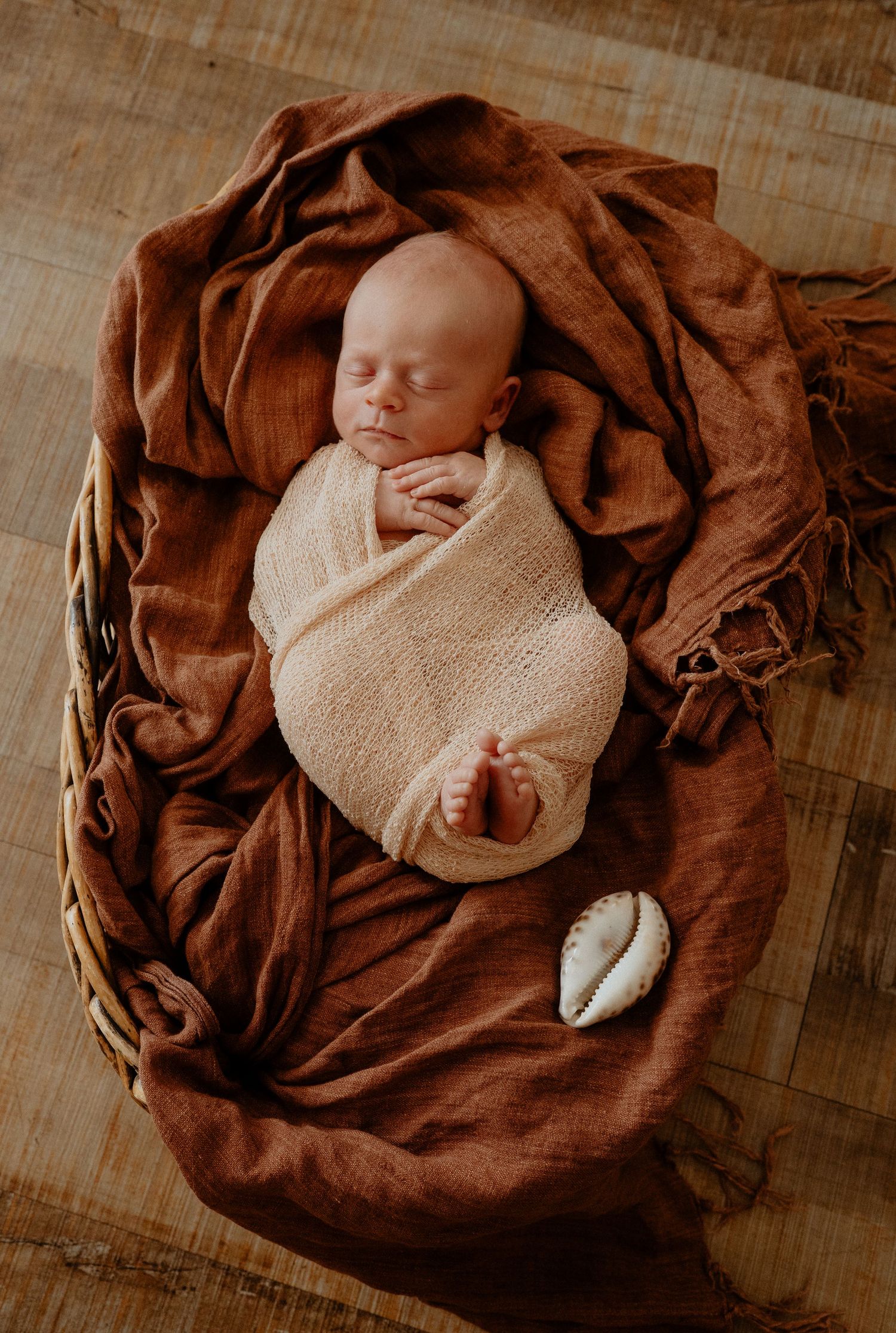 A peacefully sleeping newborn baby swaddled in cream wrap rests in a wicker basket with russet linen.