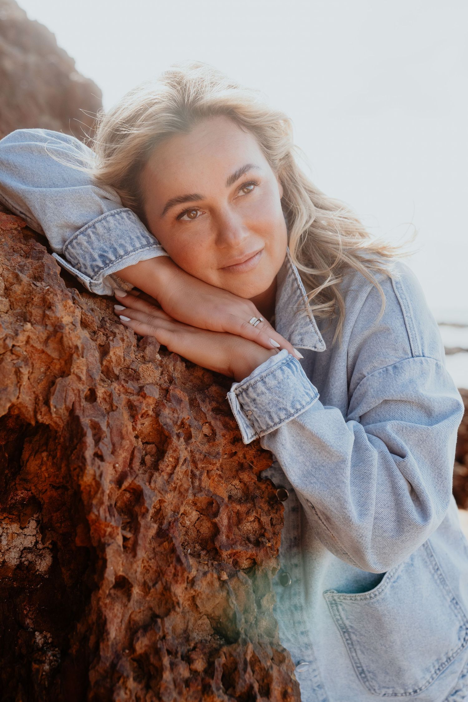 Blonde woman in denim jacket leaning on a red rock formation at the beach.