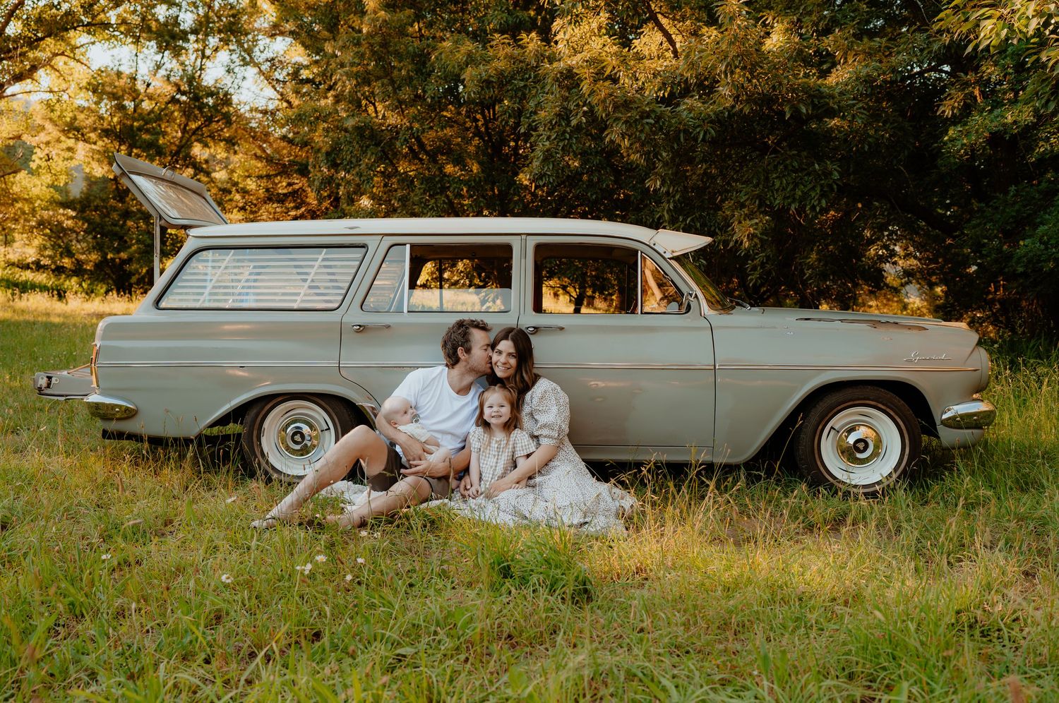 Family sitting in a grassy field beside a vintage station wagon at sunset.