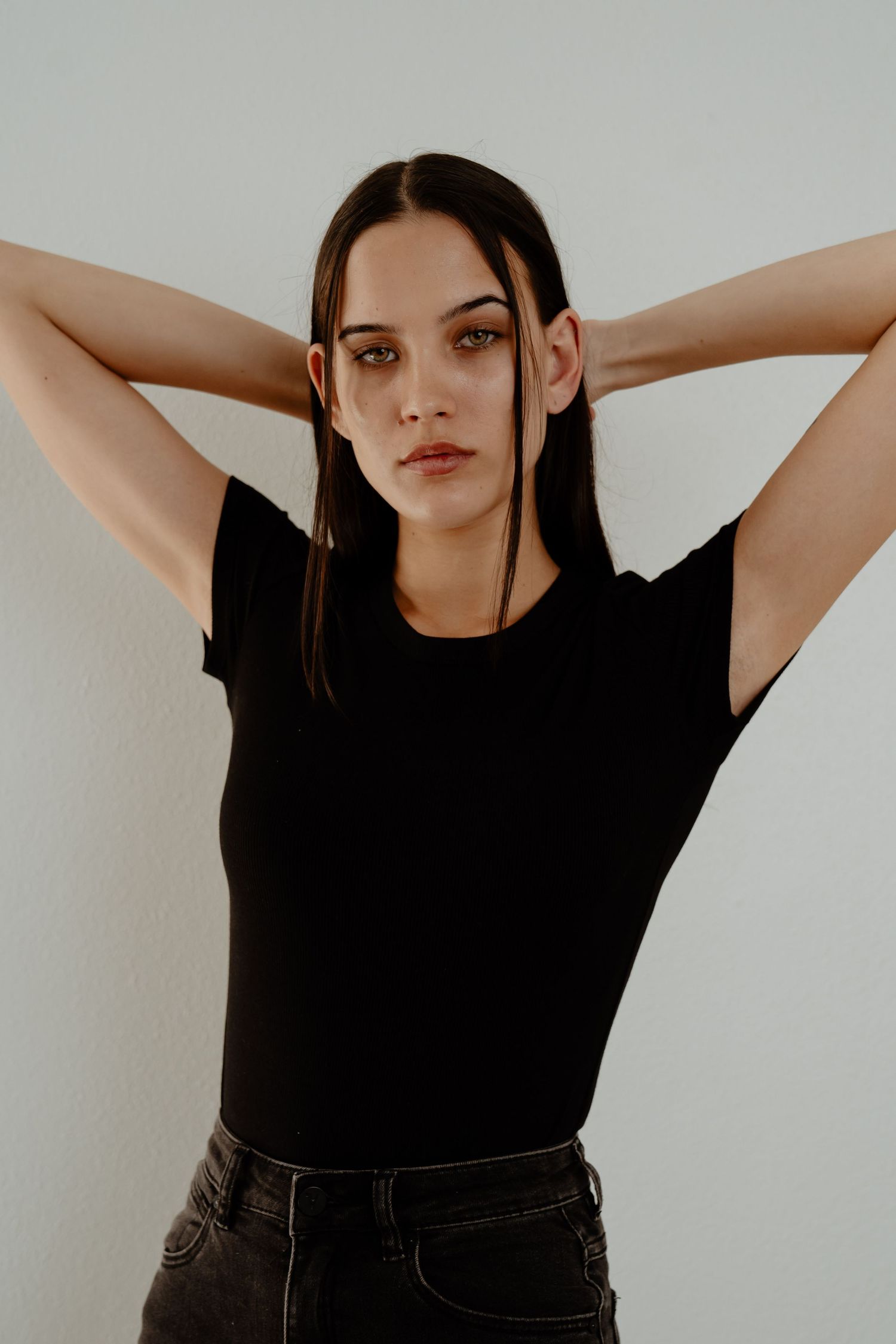 Young brunette woman in black t-shirt posing with hands behind head against a plain white background.