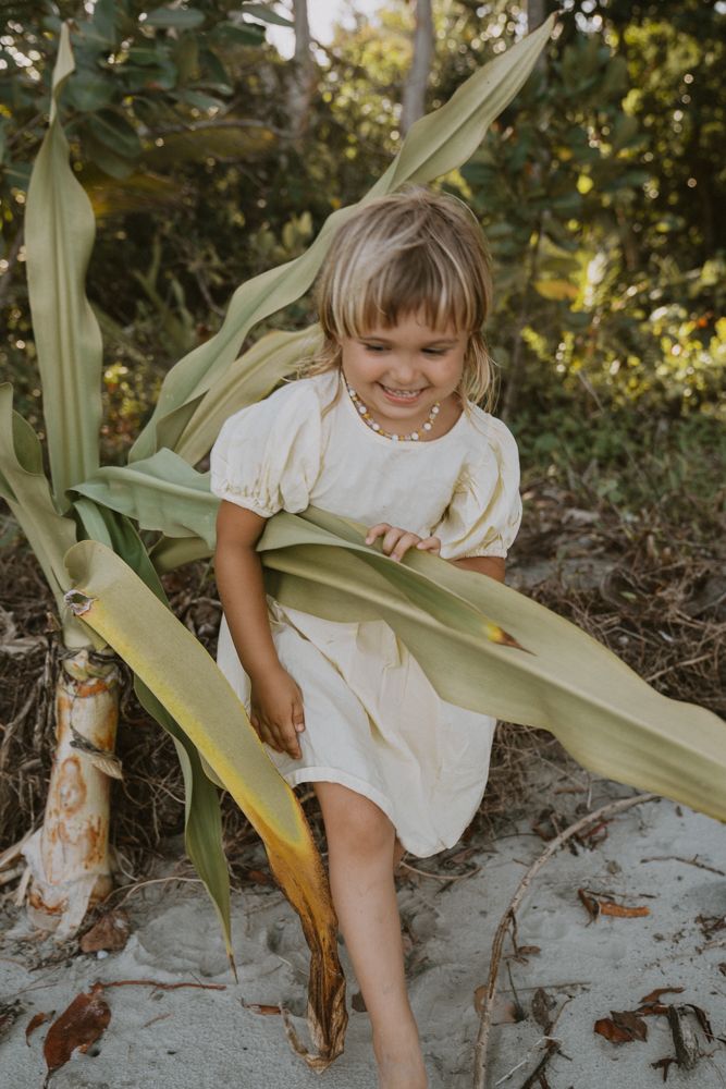 Smiling young girl in white dress sitting among large tropical green leaves on sand.