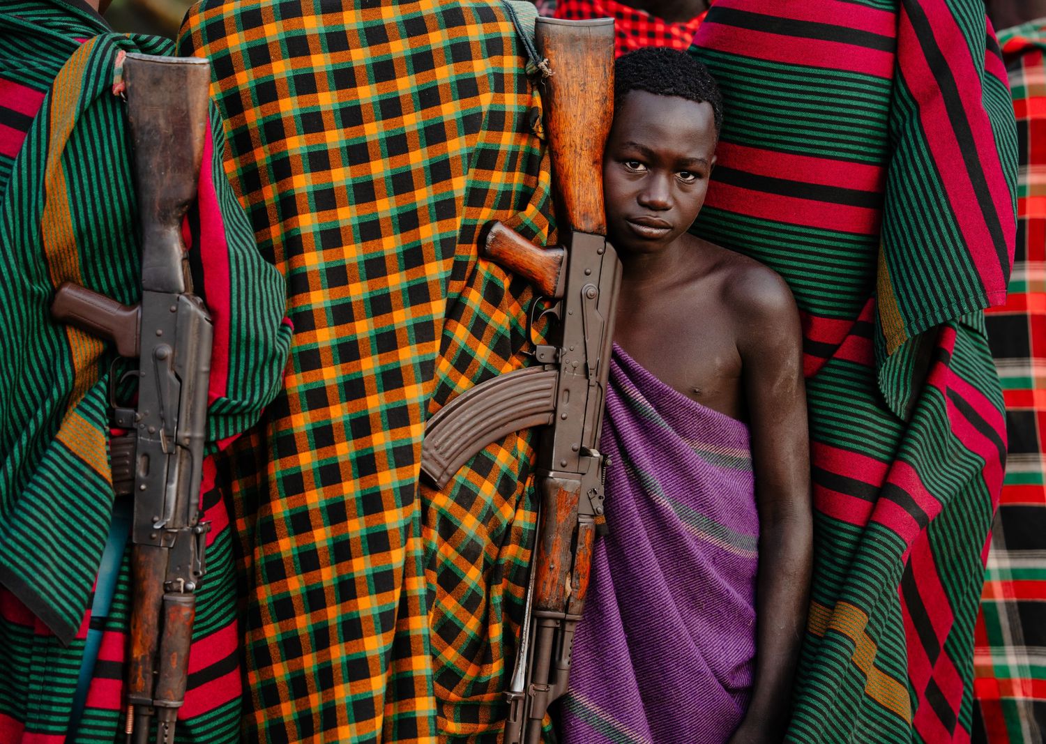 Young boy in purple cloth stands among adults in colorful traditional garments holding rifles in Africa.