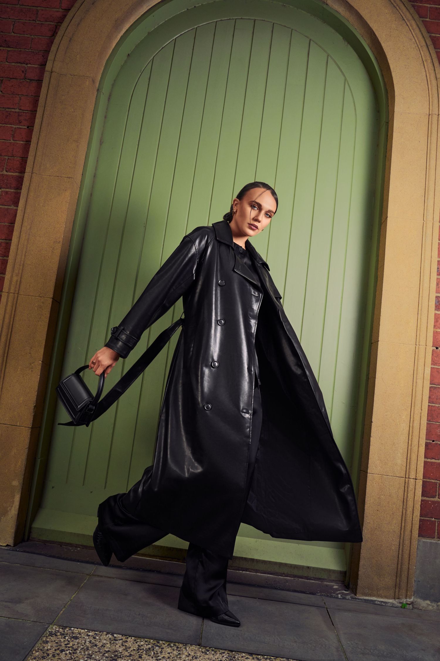 Woman in long black patent leather trench coat holding handbag by green arched door.