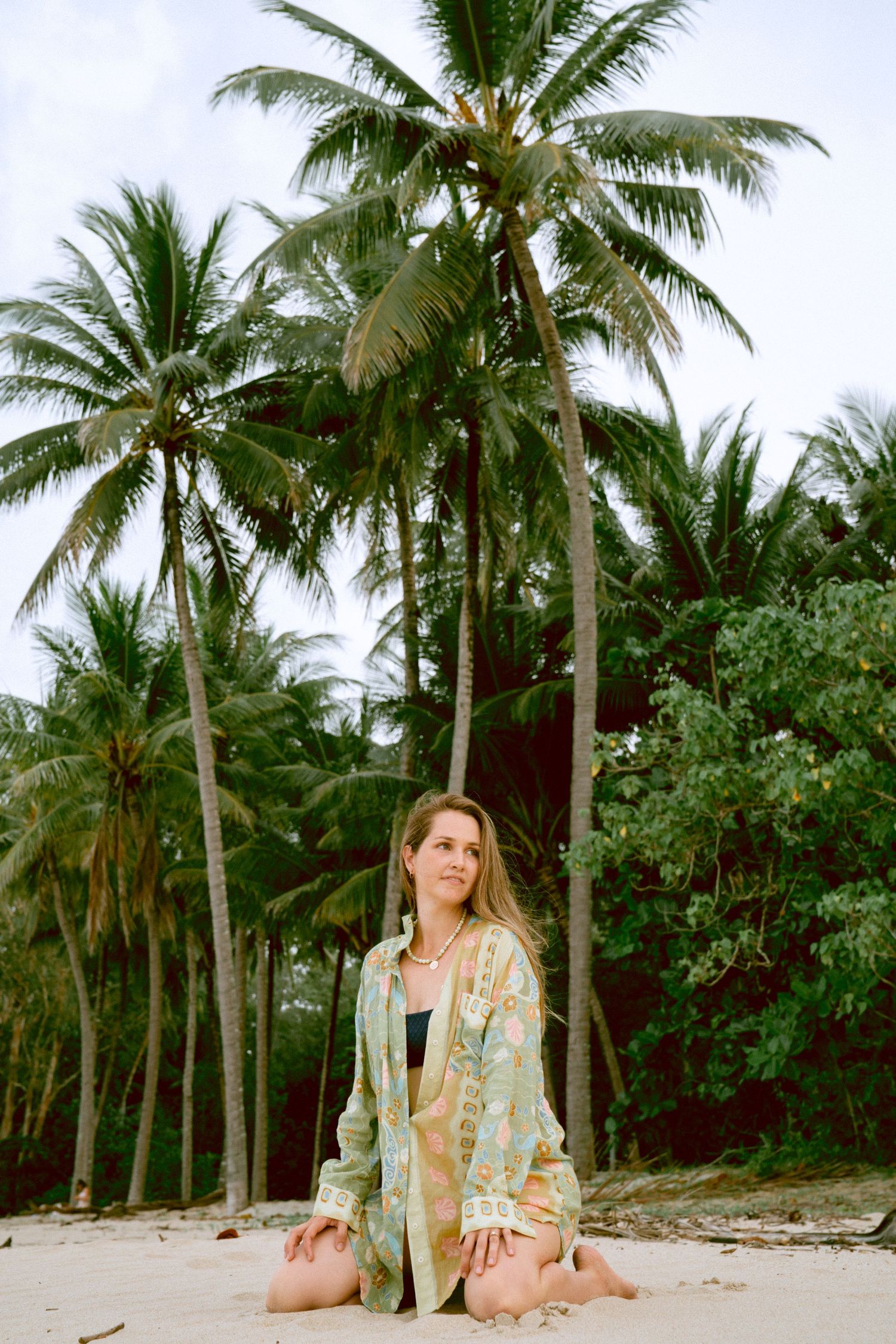 Woman in floral kimono sitting on sand surrounded by tall tropical palm trees.