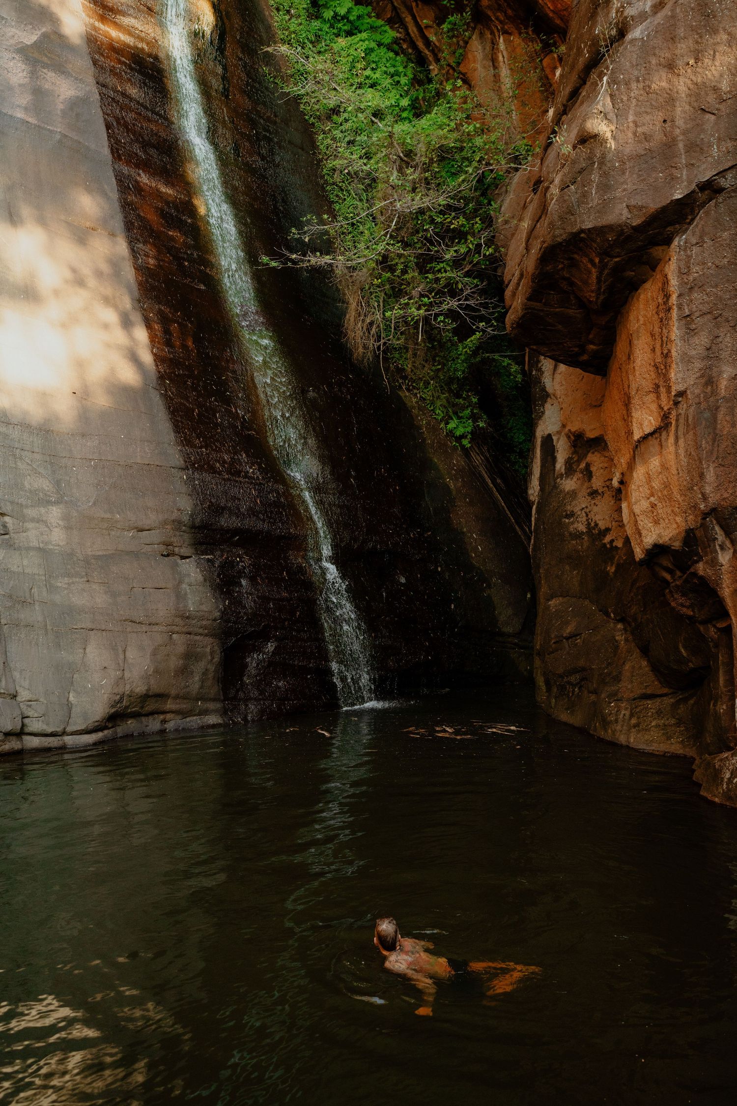 Person swimming in a tranquil rock pool beneath a stunning waterfall surrounded by sandstone cliffs.