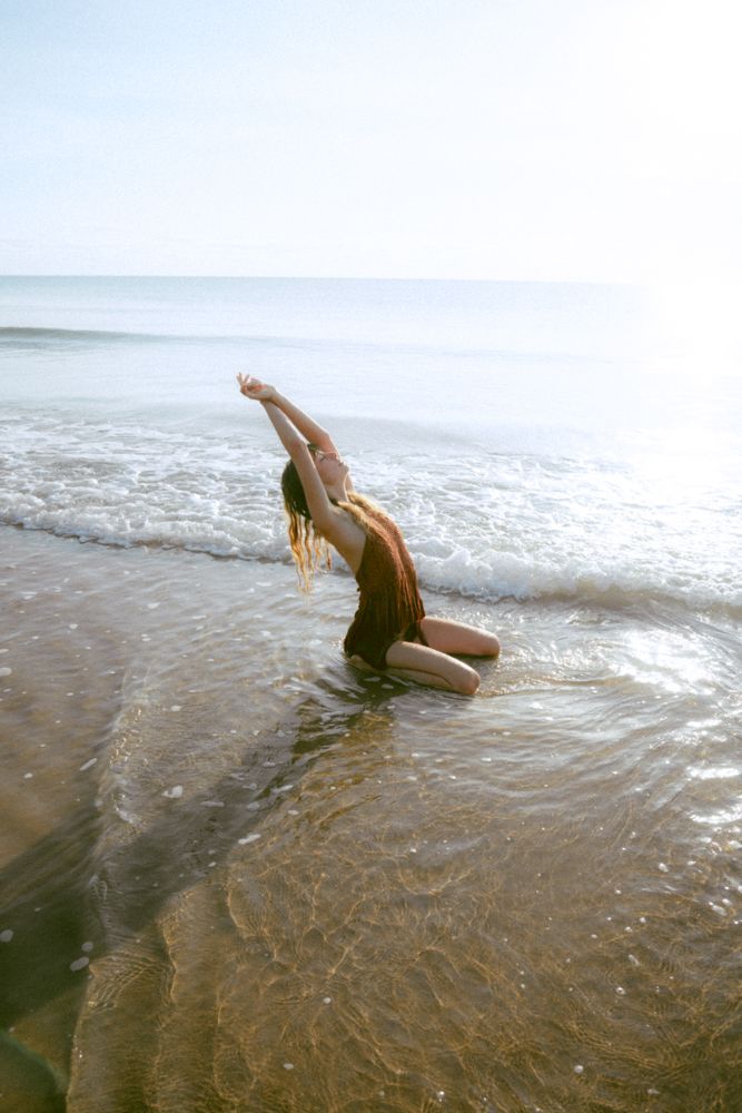 Woman kneeling in ocean waves on sandy beach, arms raised toward sky.
